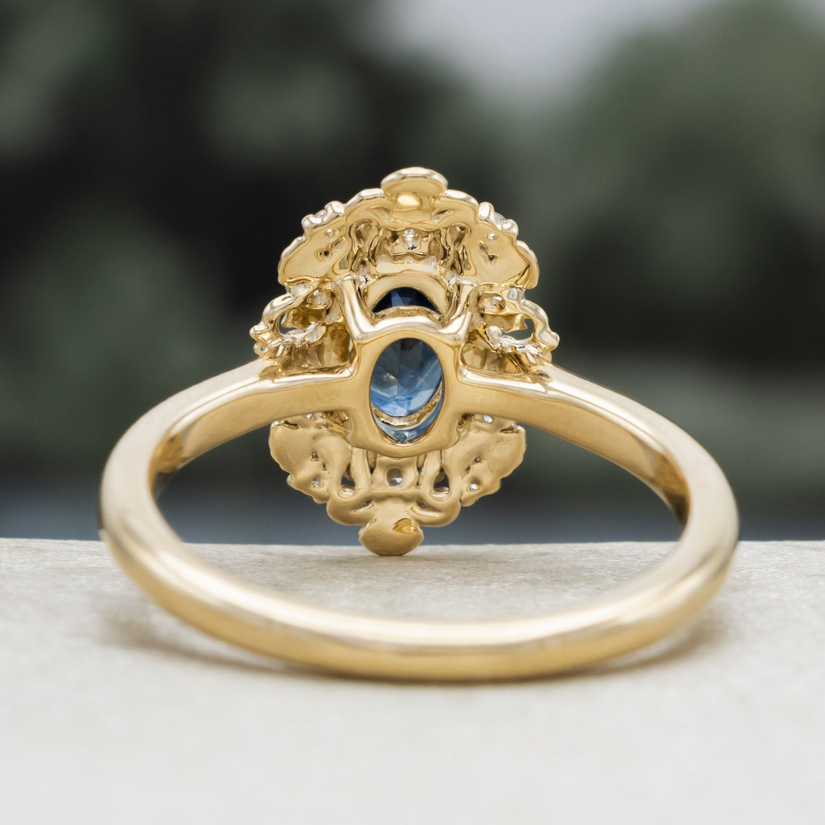 A gold ring with Montana sapphire center stone and diamond accents against a blurred green background