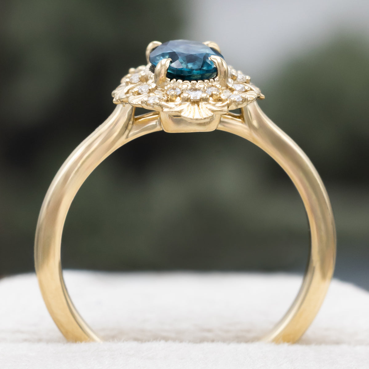 A gold ring with Montana sapphire center stone and diamond accents against a blurred green background