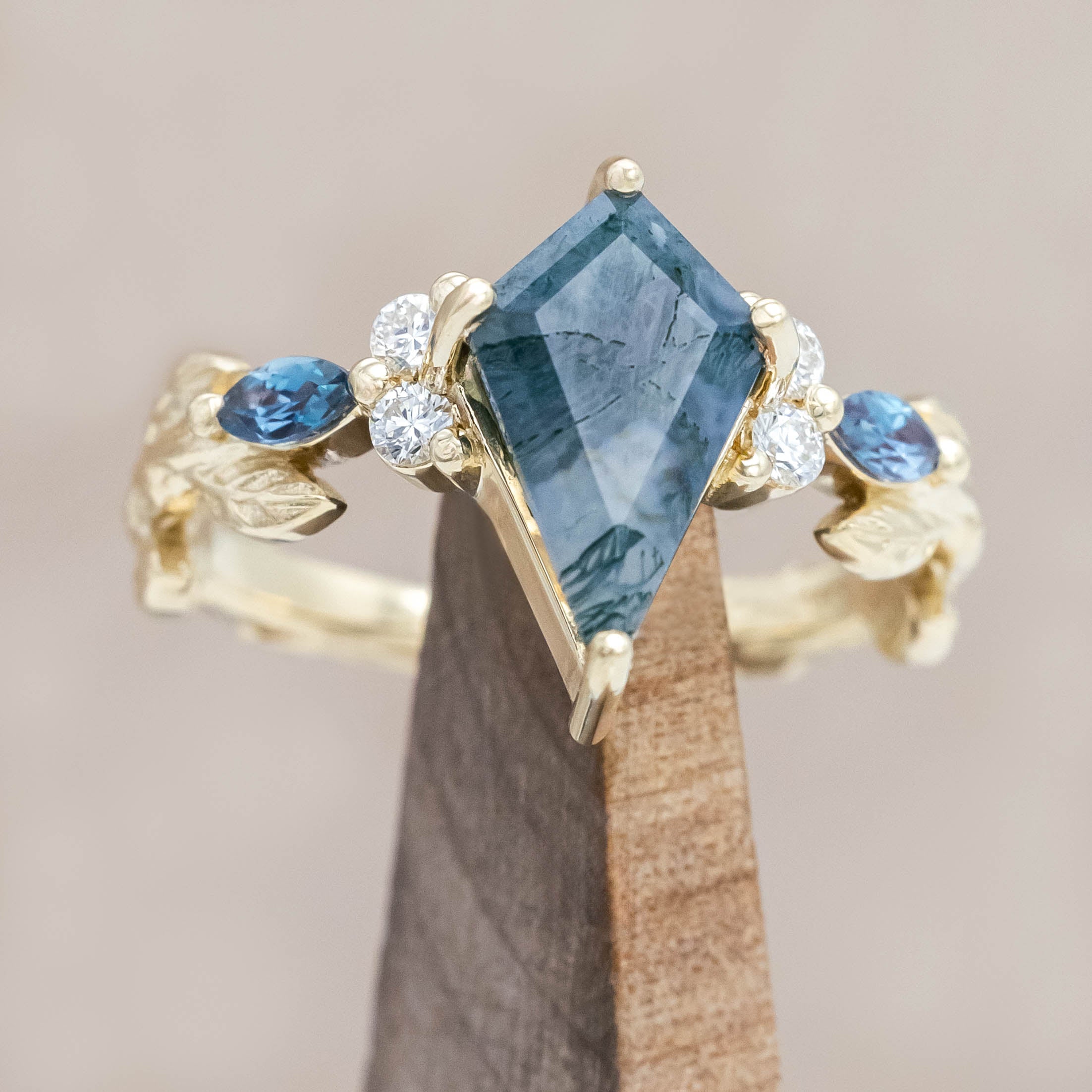 Gold ring with moss agate center stone with alexandrite & diamond accents on a wooden stand against a neutral background