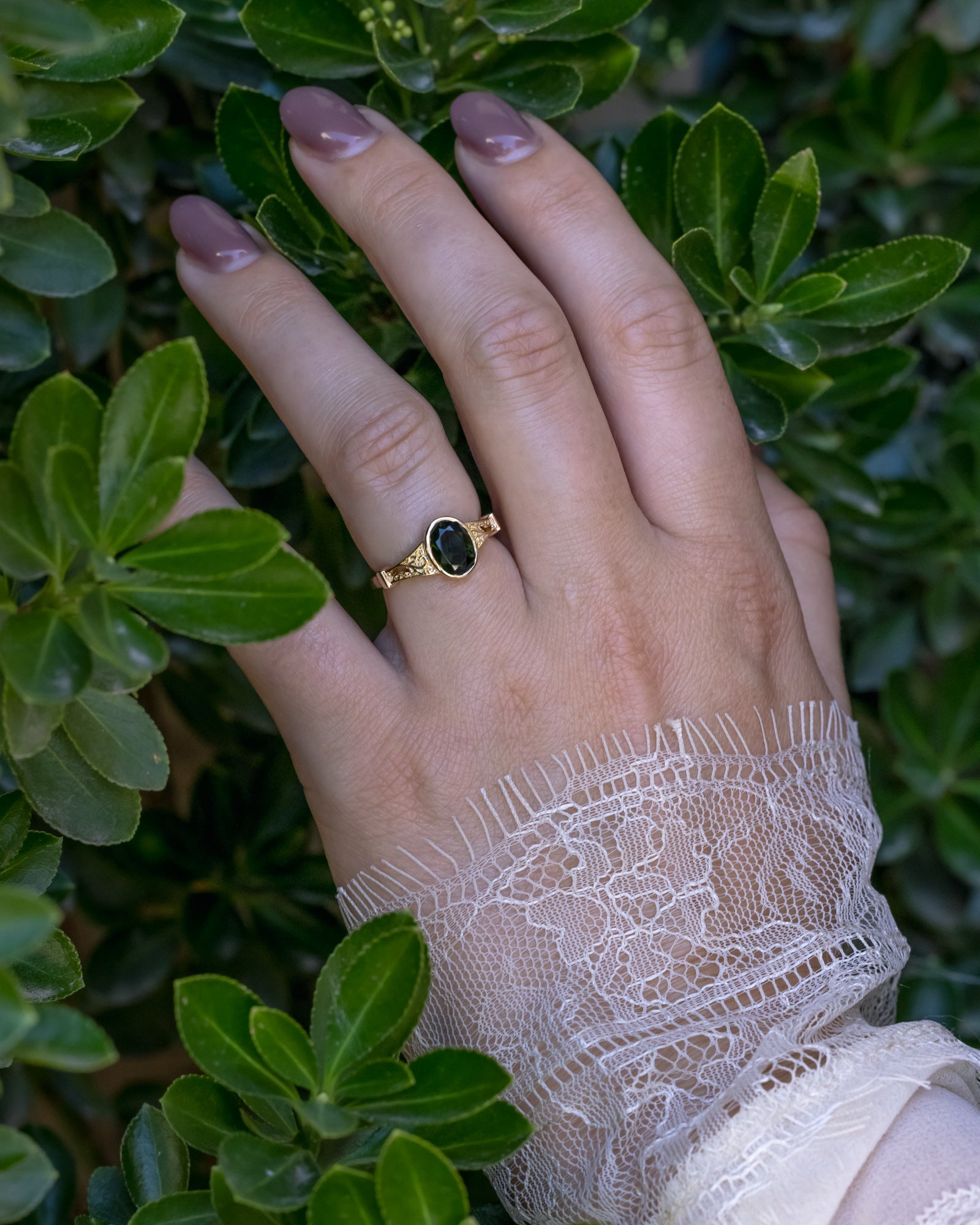 Hand wearing a gold ring with a dark green tourmaline against a green leafy background