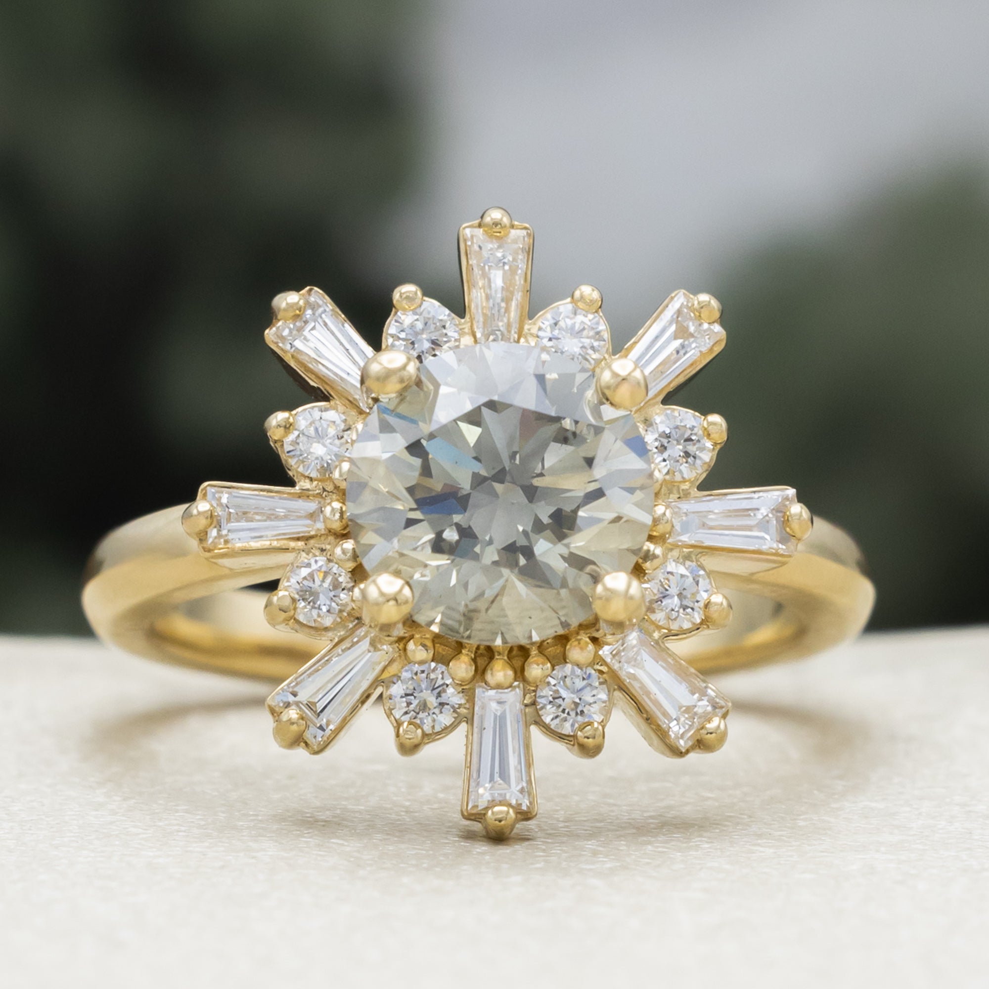 A yellow gold ring with salt & pepper diamond center stone and diamond accents on a blurred green background