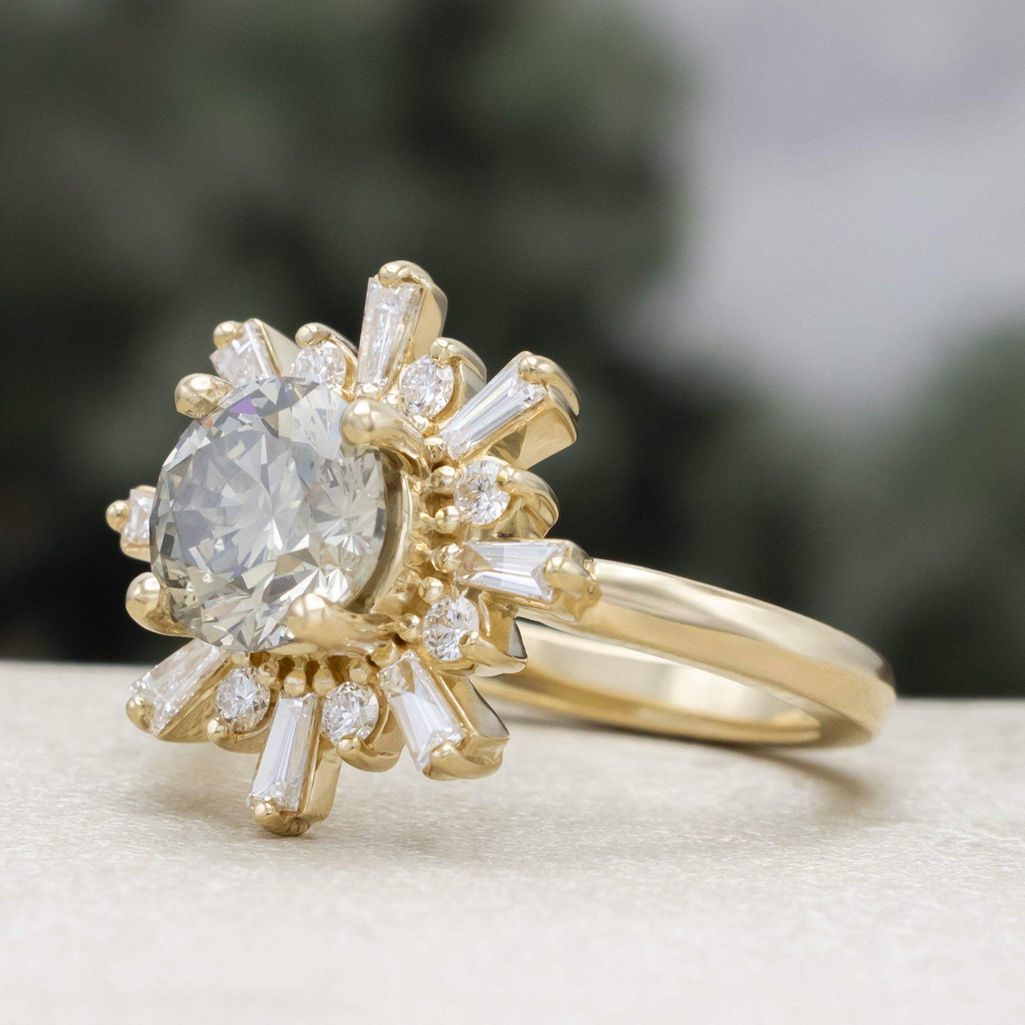 A yellow gold ring with salt & pepper diamond center stone and diamond accents on a blurred green background