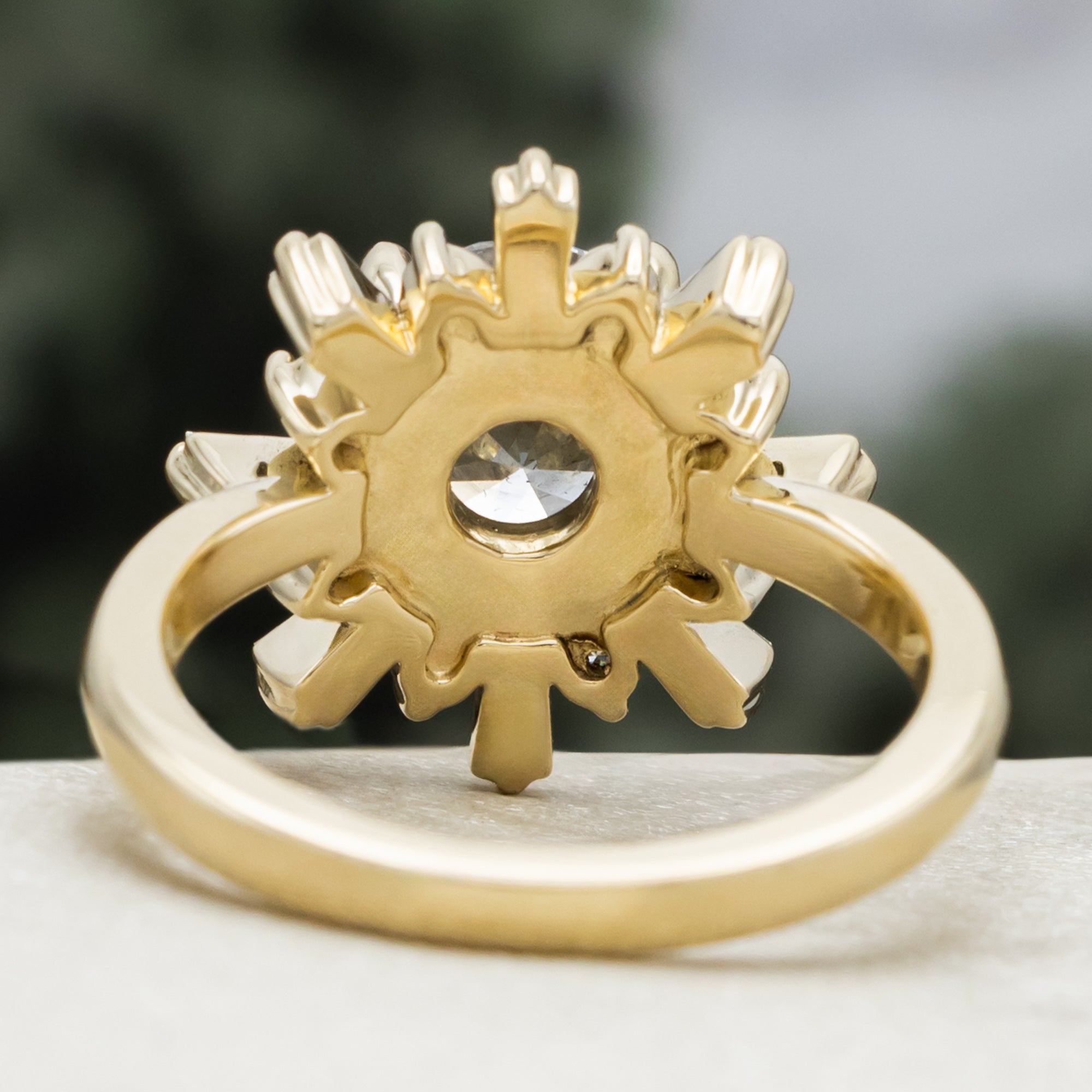 A yellow gold ring with salt & pepper diamond center stone and diamond accents on a blurred green background