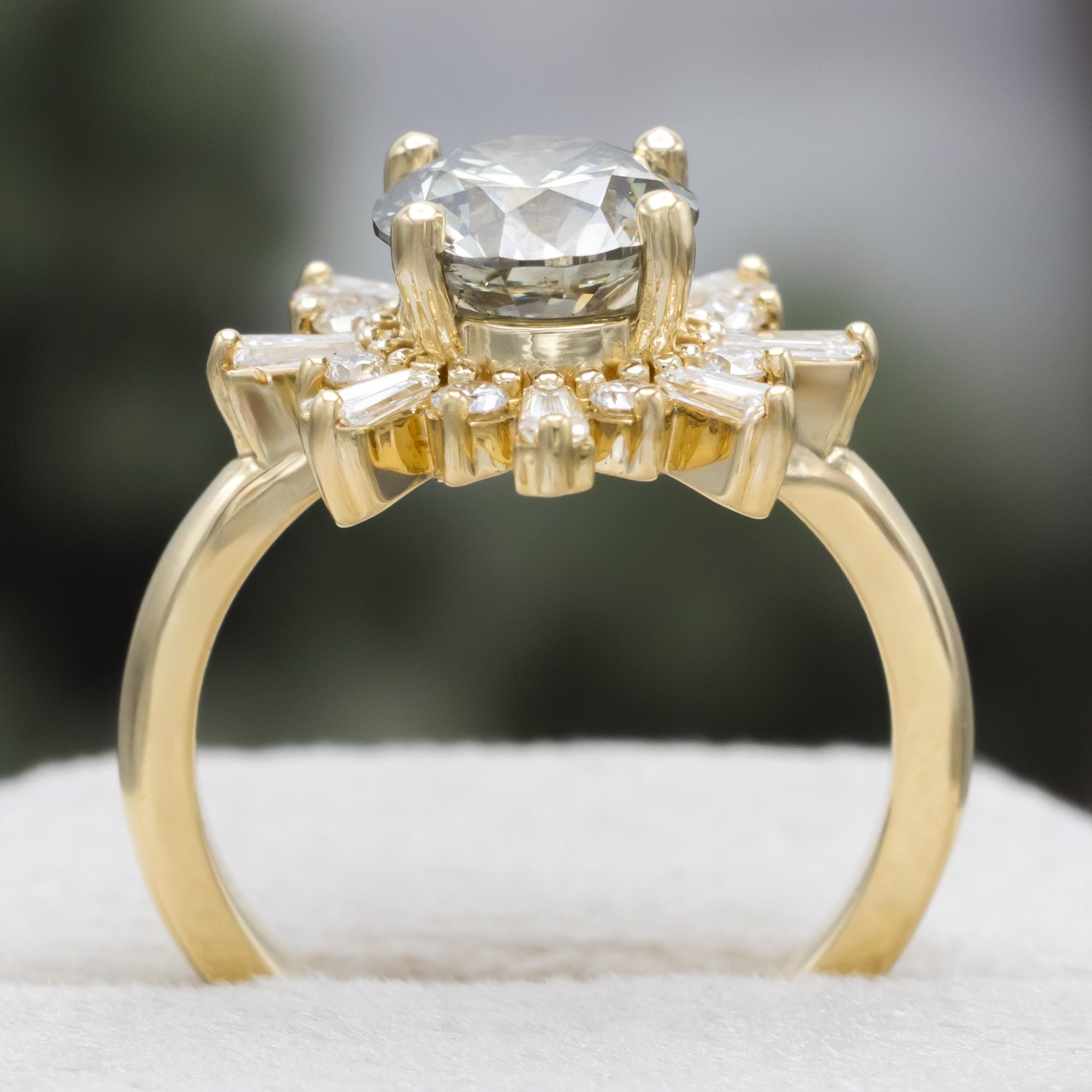 A yellow gold ring with salt & pepper diamond center stone and diamond accents on a blurred green background