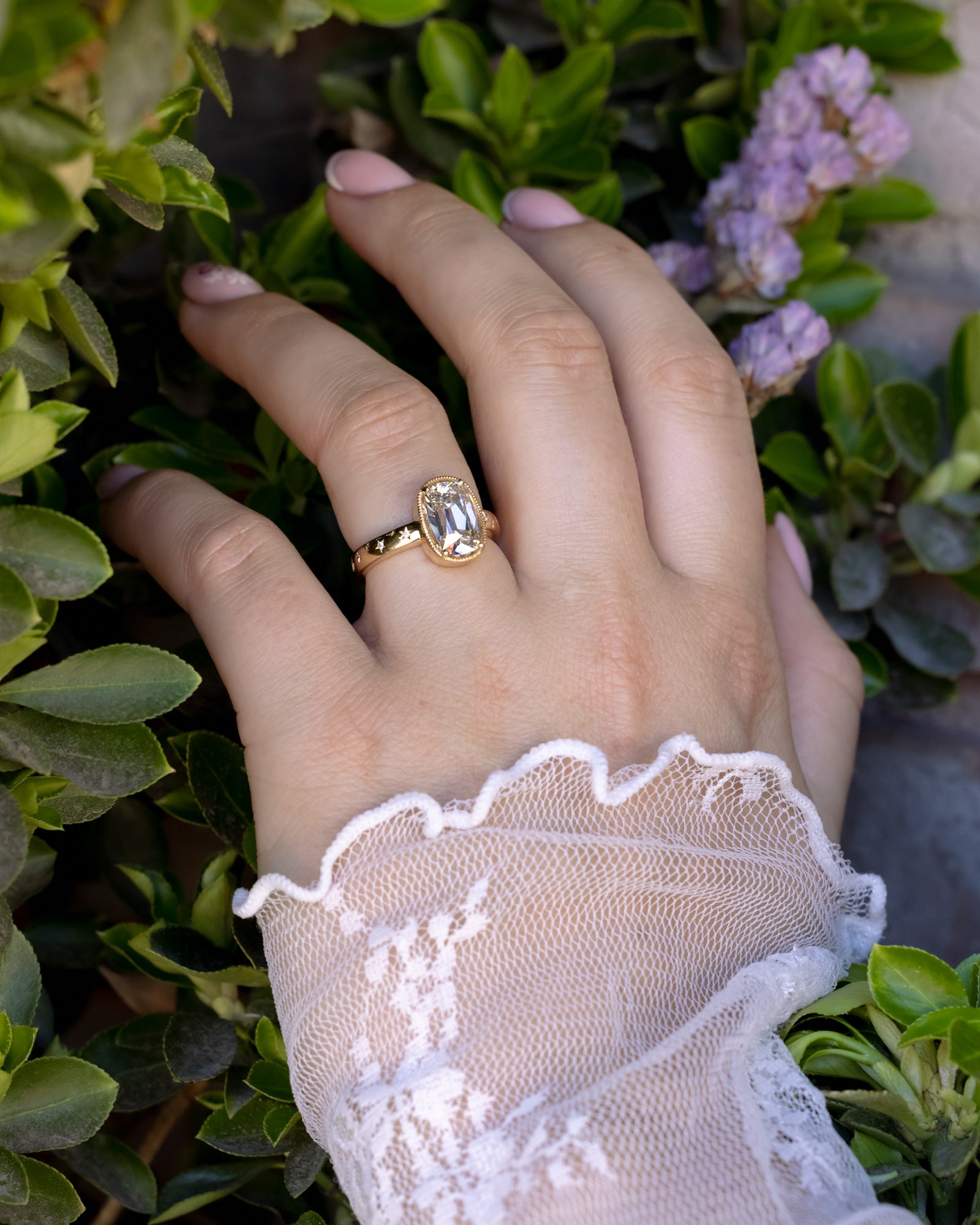 Hand wearing a gold ring with a lab diamond, covered by a lace sleeve, against a green leafy background.