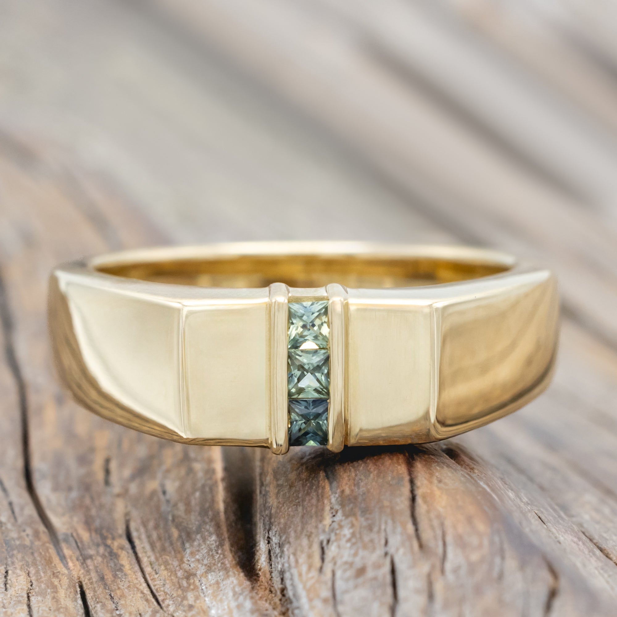 A polished yellow gold ring with a central channel set featuring light blue and green sapphires on a wooden surface.