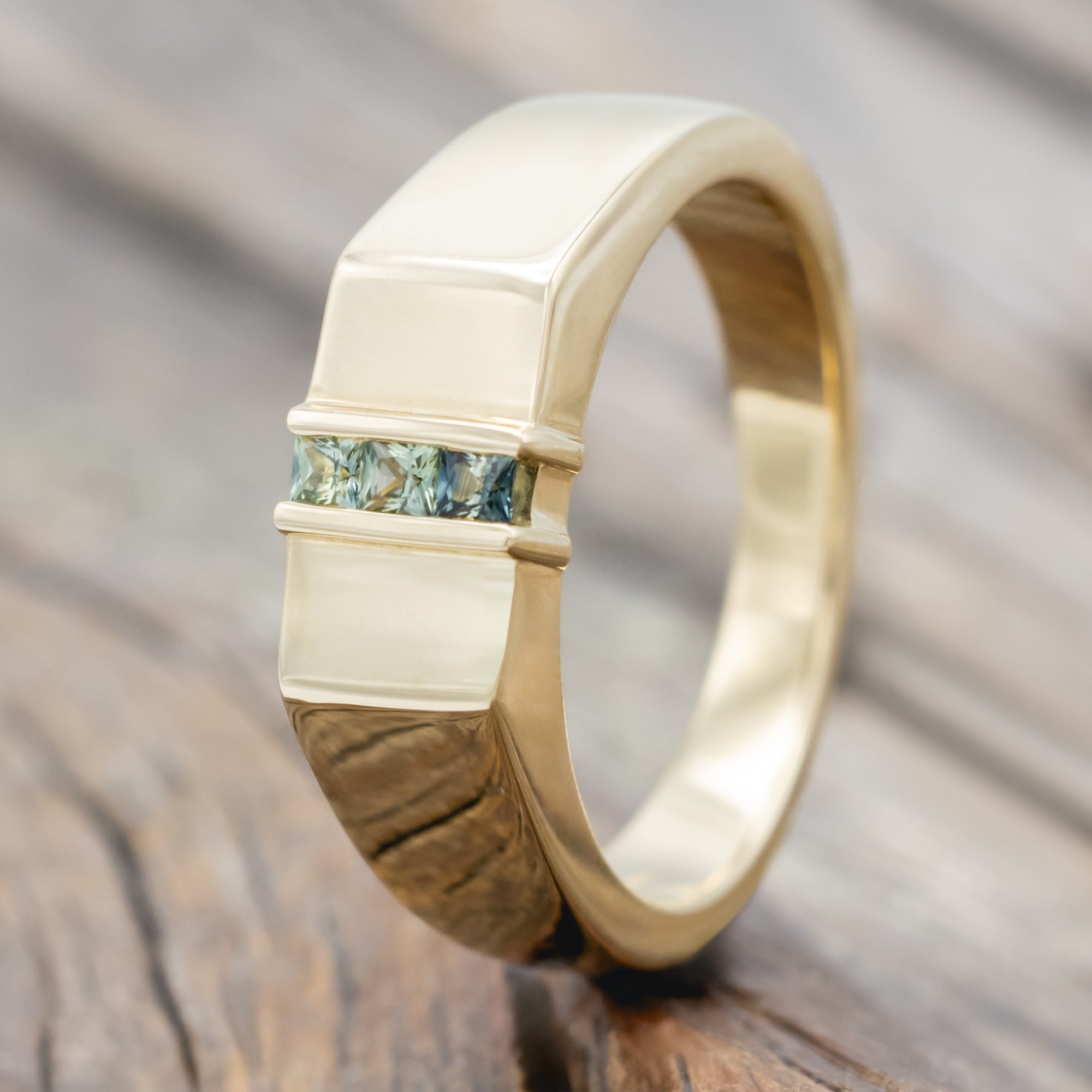 A polished yellow gold ring with a central channel set featuring light blue and green sapphires on a wooden surface.