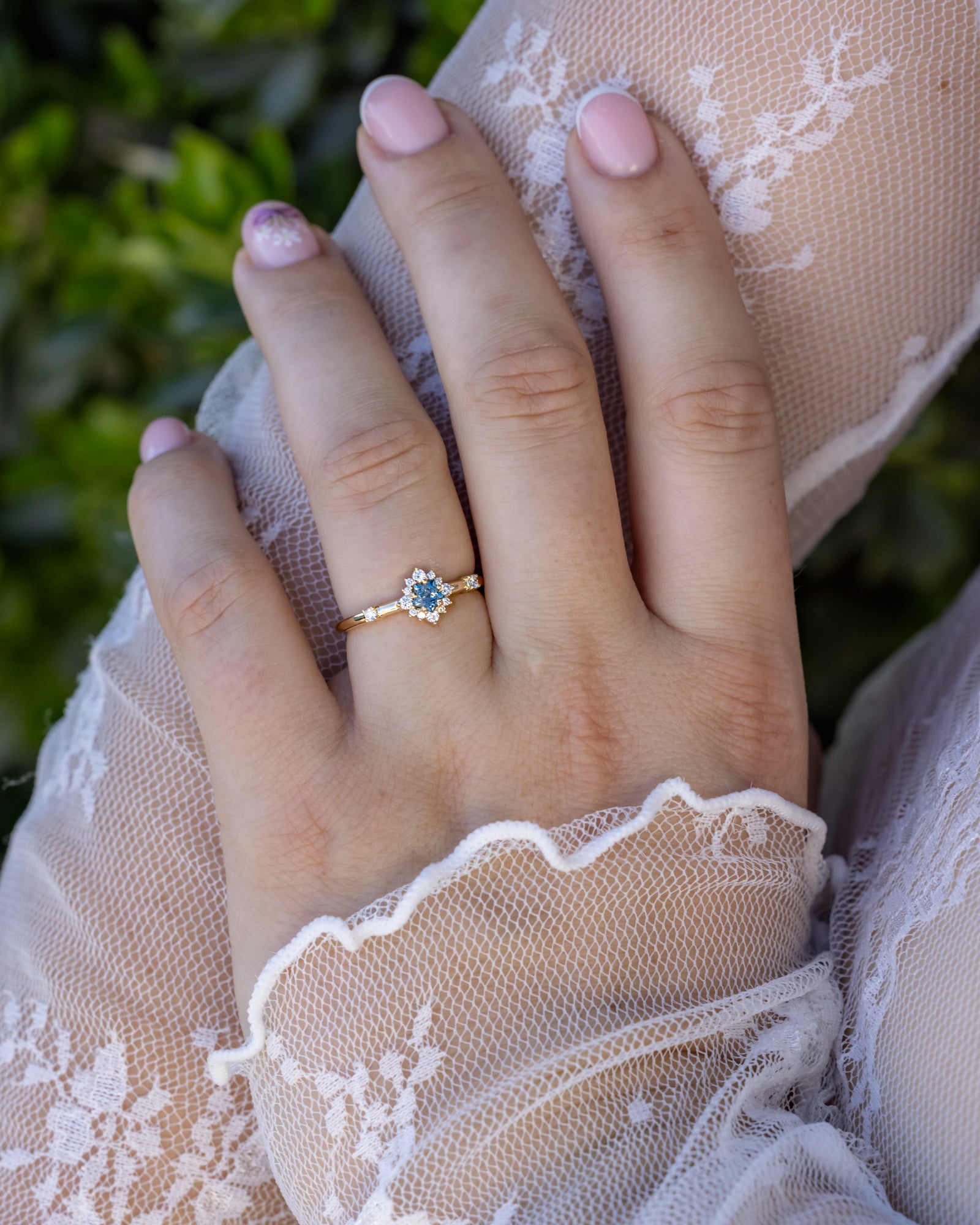 A hand wearing a gold ring with a blue sapphire, wearing a lace sleeve against a blurred green background