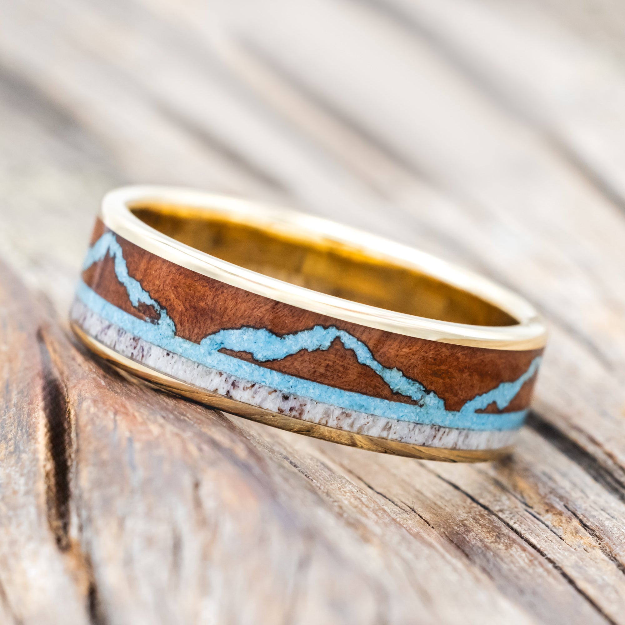 Yellow gold ring with whiskey barrel, antler, and turquoise mountains on a wooden surface