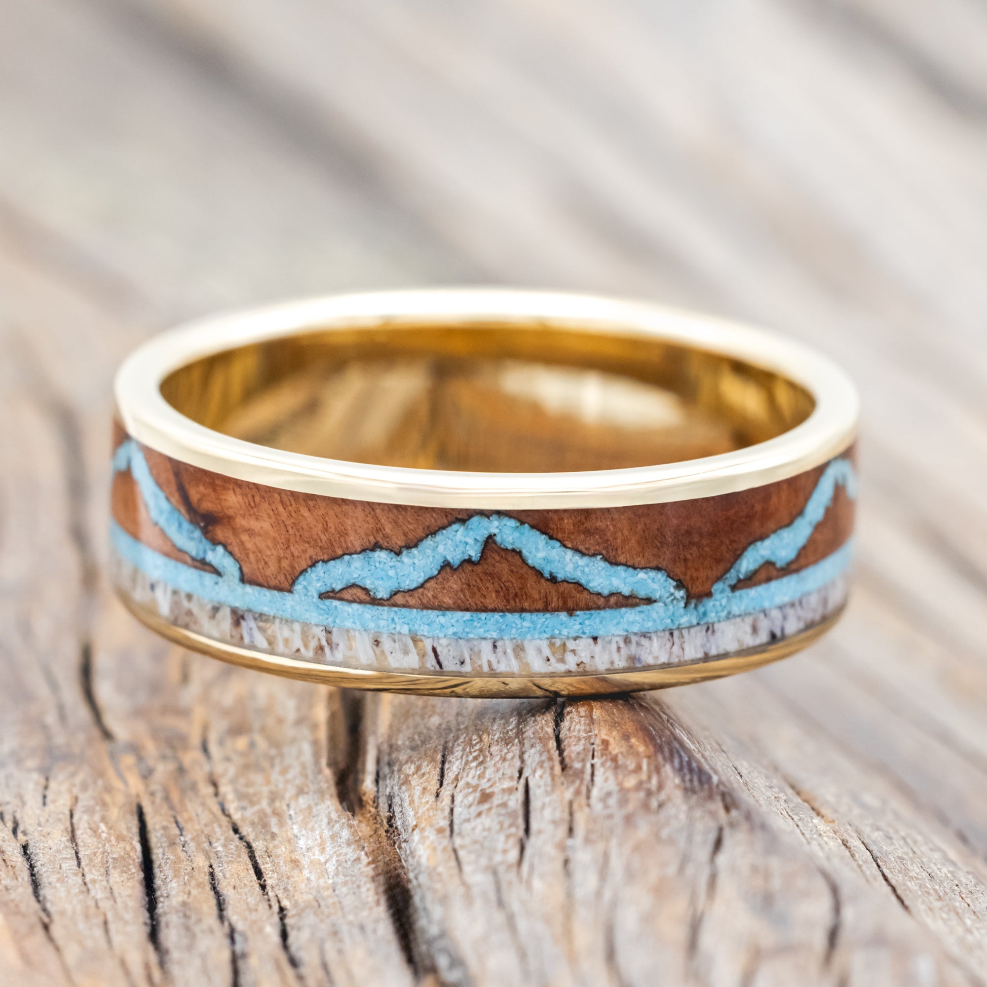Yellow gold ring with whiskey barrel, antler, and turquoise mountains on a wooden surface