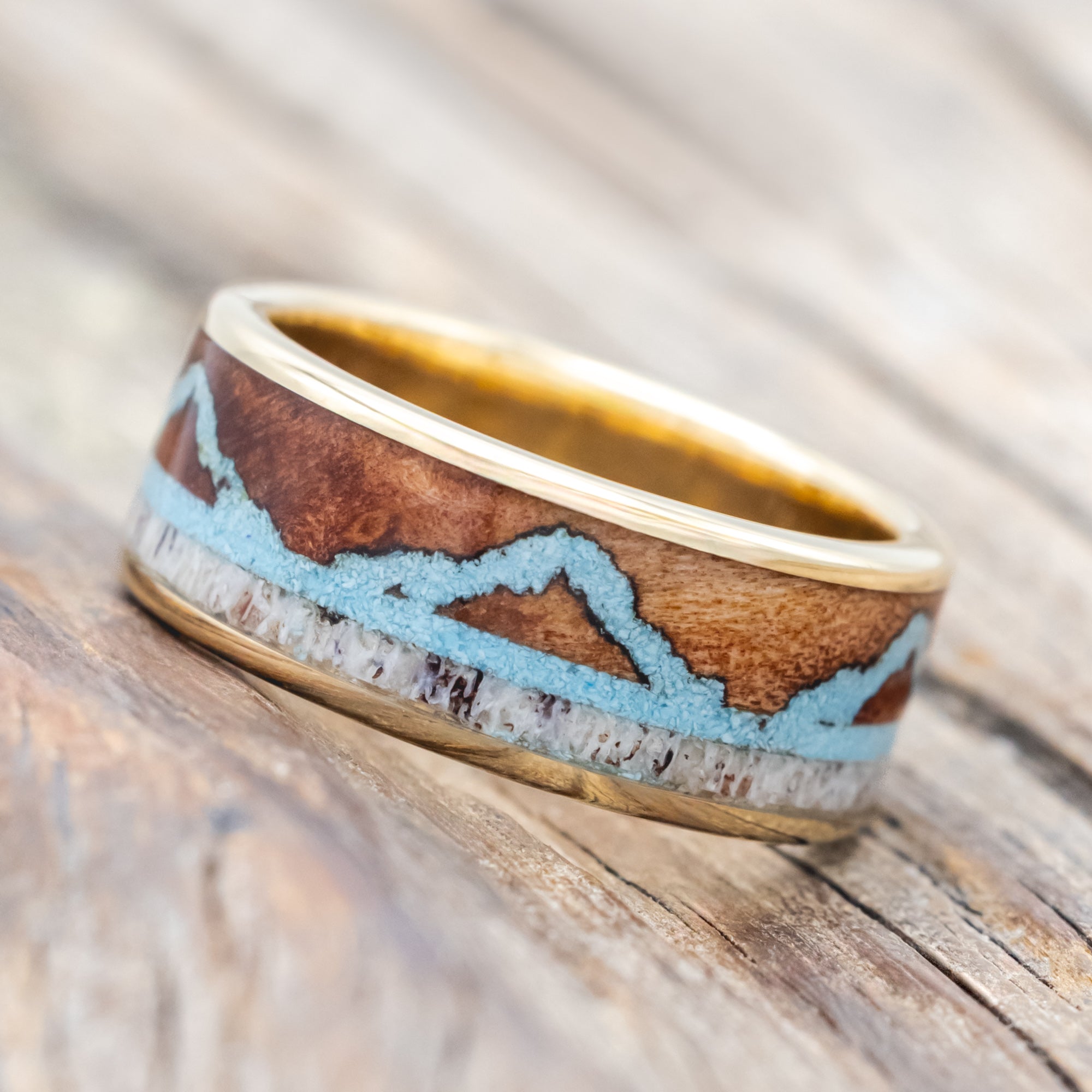 Yellow gold ring with whiskey barrel, antler, and turquoise mountains on a wooden surface