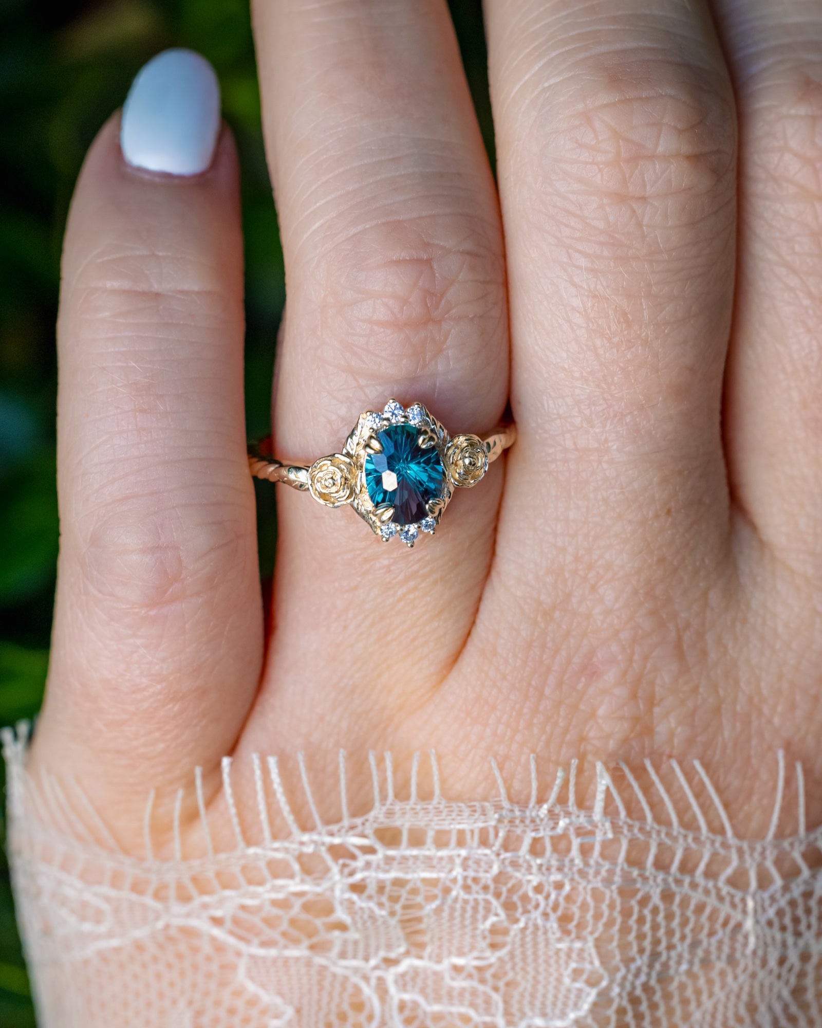 On hand photo of floral-inspired ring with alexandrite centerstone and diamond accents against blurred green background