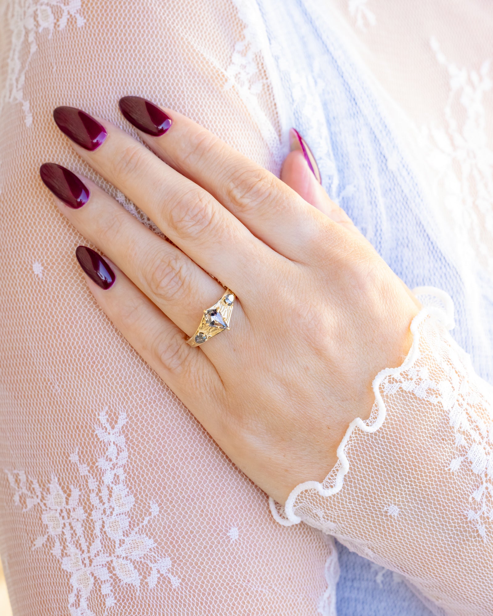 Hand with maroon nail polish wearing a salt & pepper diamond gold ring on a light background