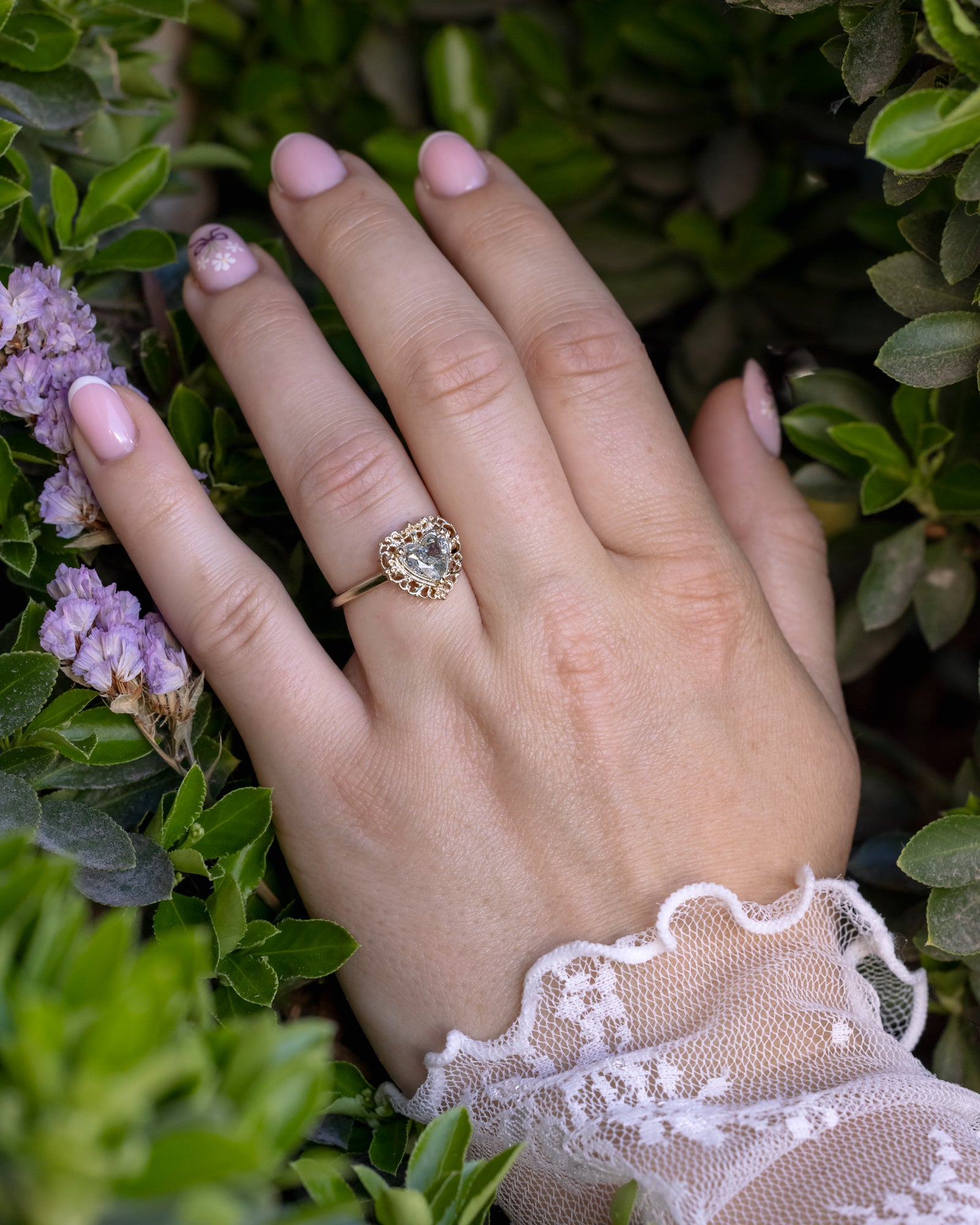 Hand wearing a gold ring with a heart-shaped salt & pepper diamond, surrounded by green foliage and purple flowers.