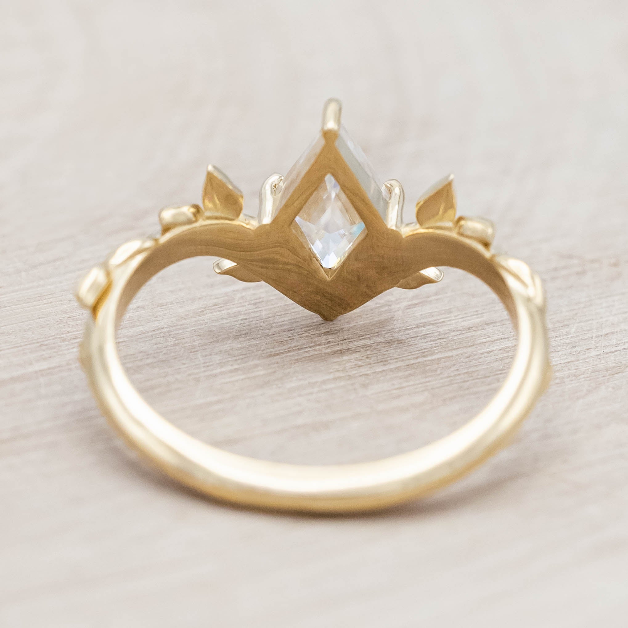 Gold vine-style ring with a lozenge Moissanite on a wooden stand against a neutral background