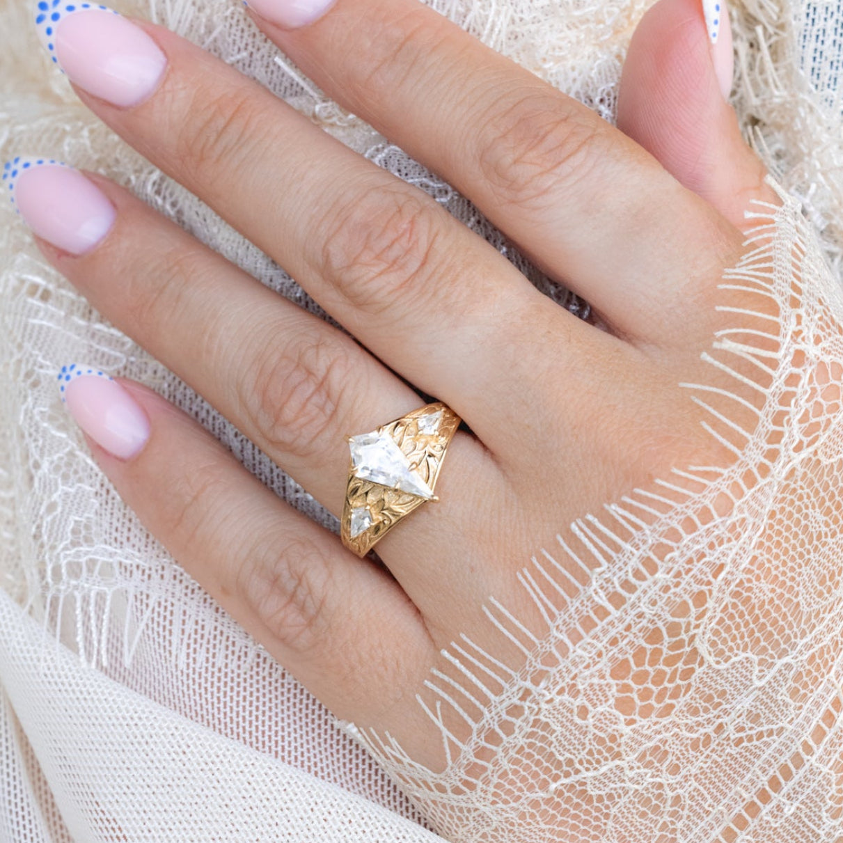 Close-up of a hand wearing a gold ring with Moissanite on a white lace fabric background.