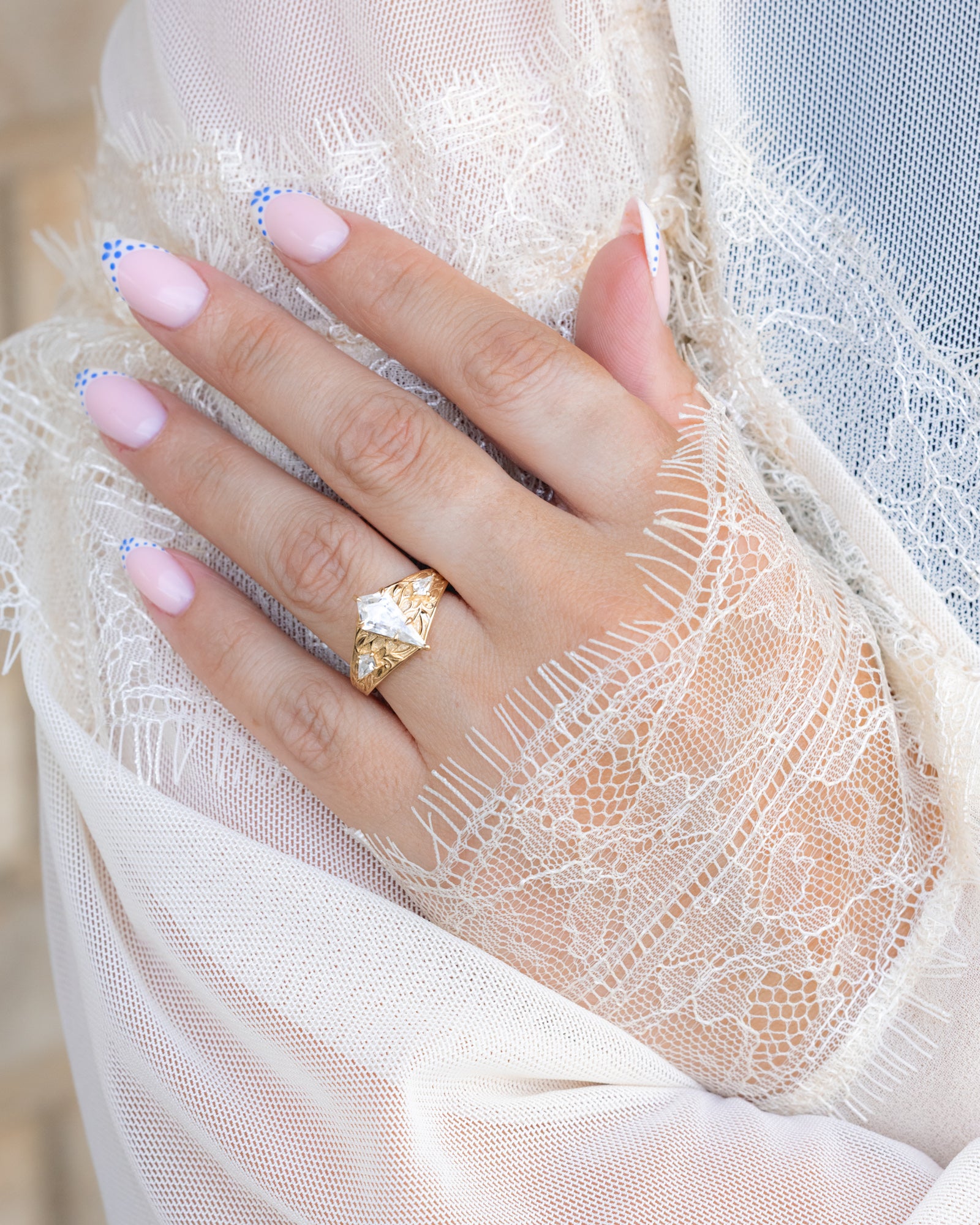 Close-up of a hand wearing a gold ring with Moissanite on a white lace fabric background.