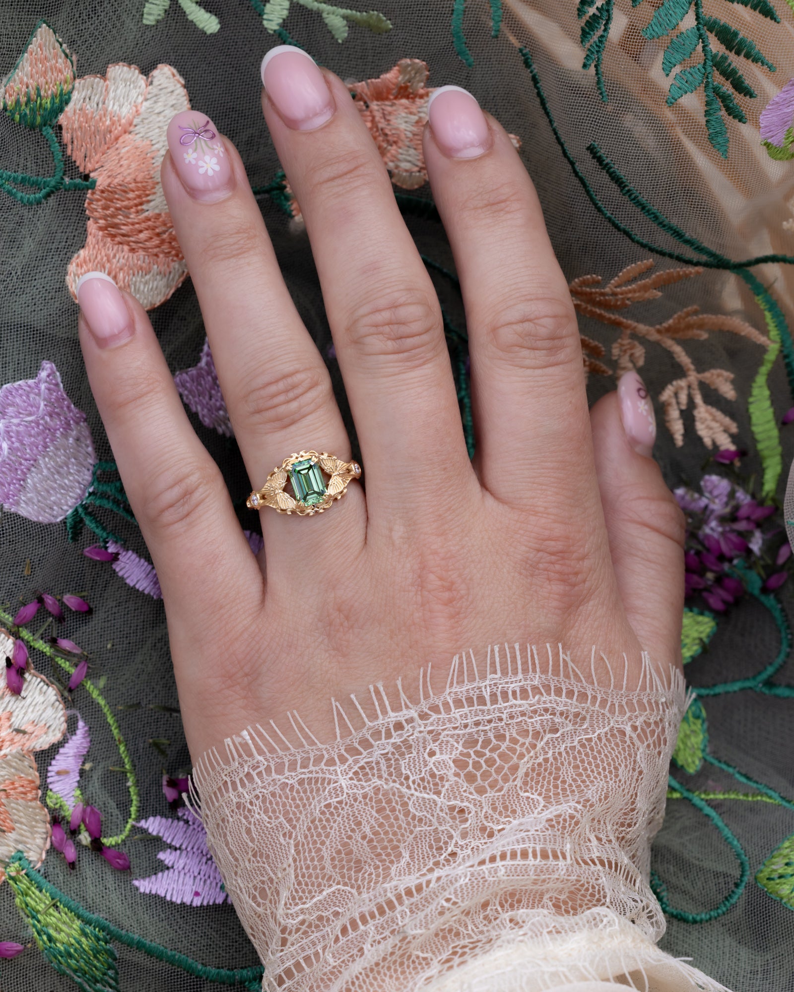 Hand wearing a gold ring with a green lab diamond on an embroidered fabric background