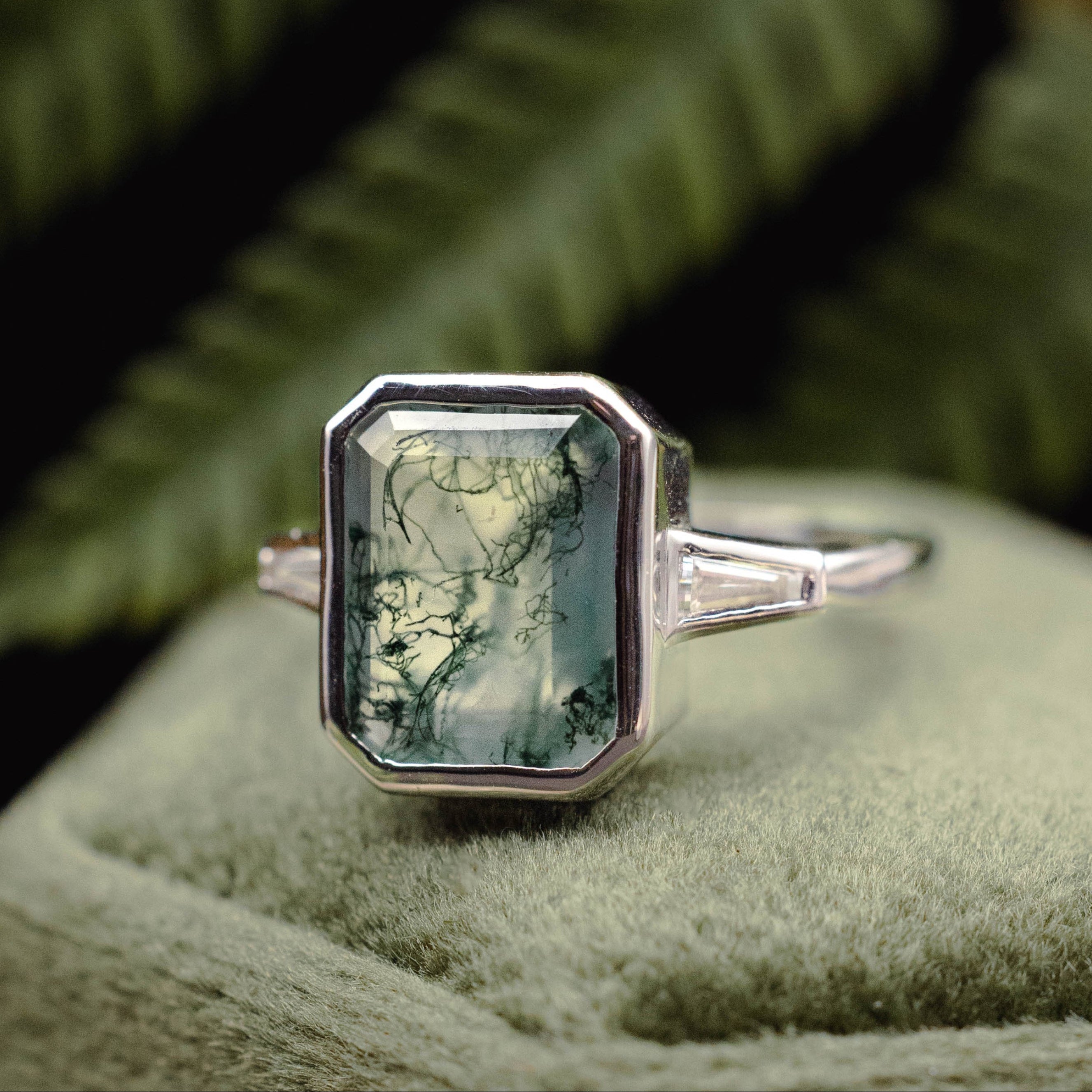 White gold ring with moss agate on a green cushion with ferns in the background