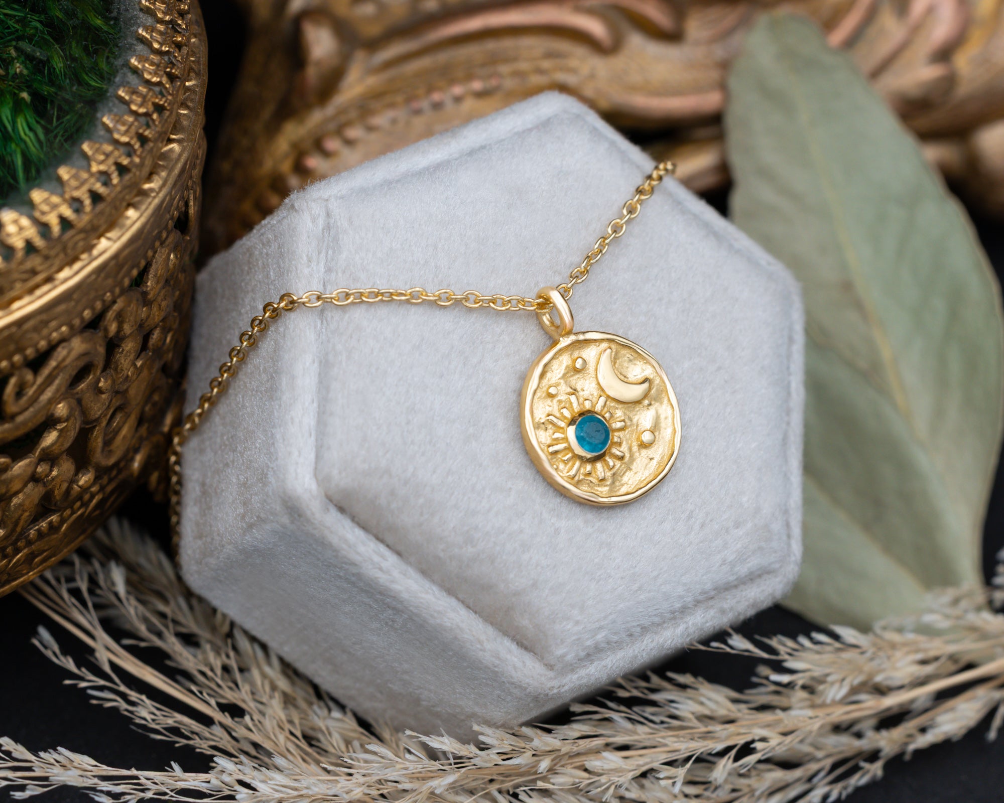 A gold pendant necklace with a crescent moon and sun design and a small blue stone, displayed on a hexagonal white jewelry box.