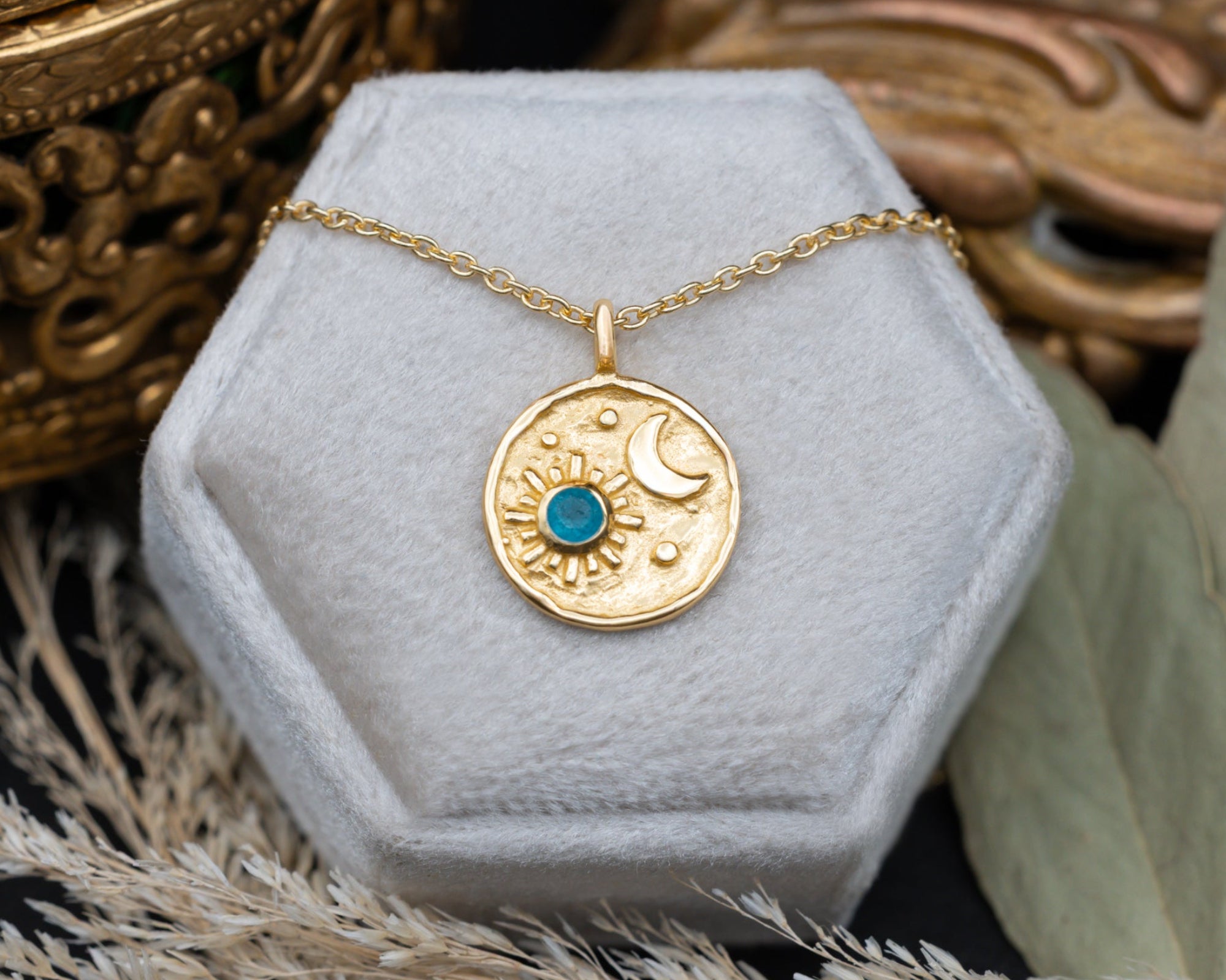 A gold pendant necklace with a crescent moon and sun design and a small blue stone, displayed on a hexagonal white jewelry box.