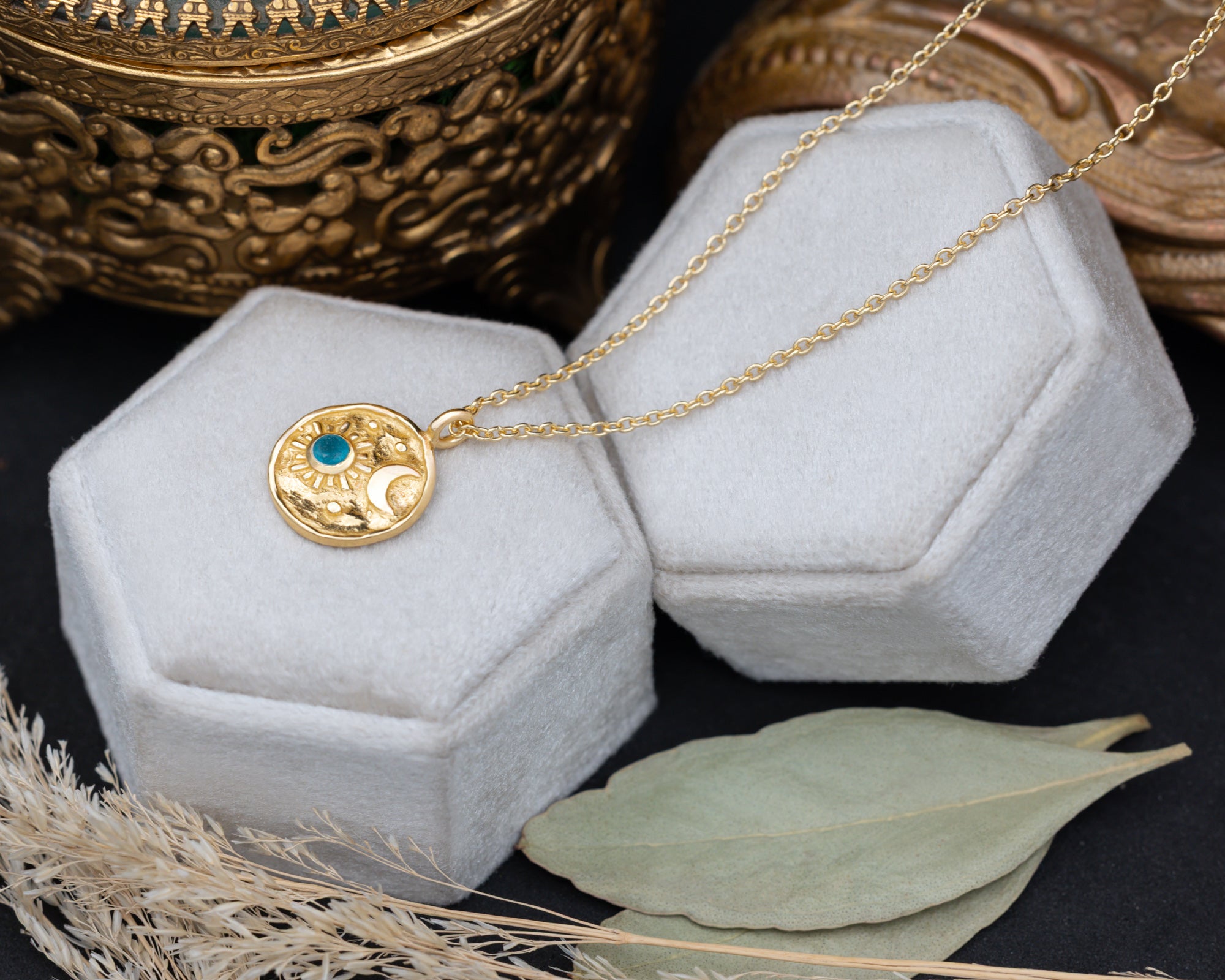 A gold pendant necklace with a crescent moon and sun design and a small blue stone, displayed on a hexagonal white jewelry box.