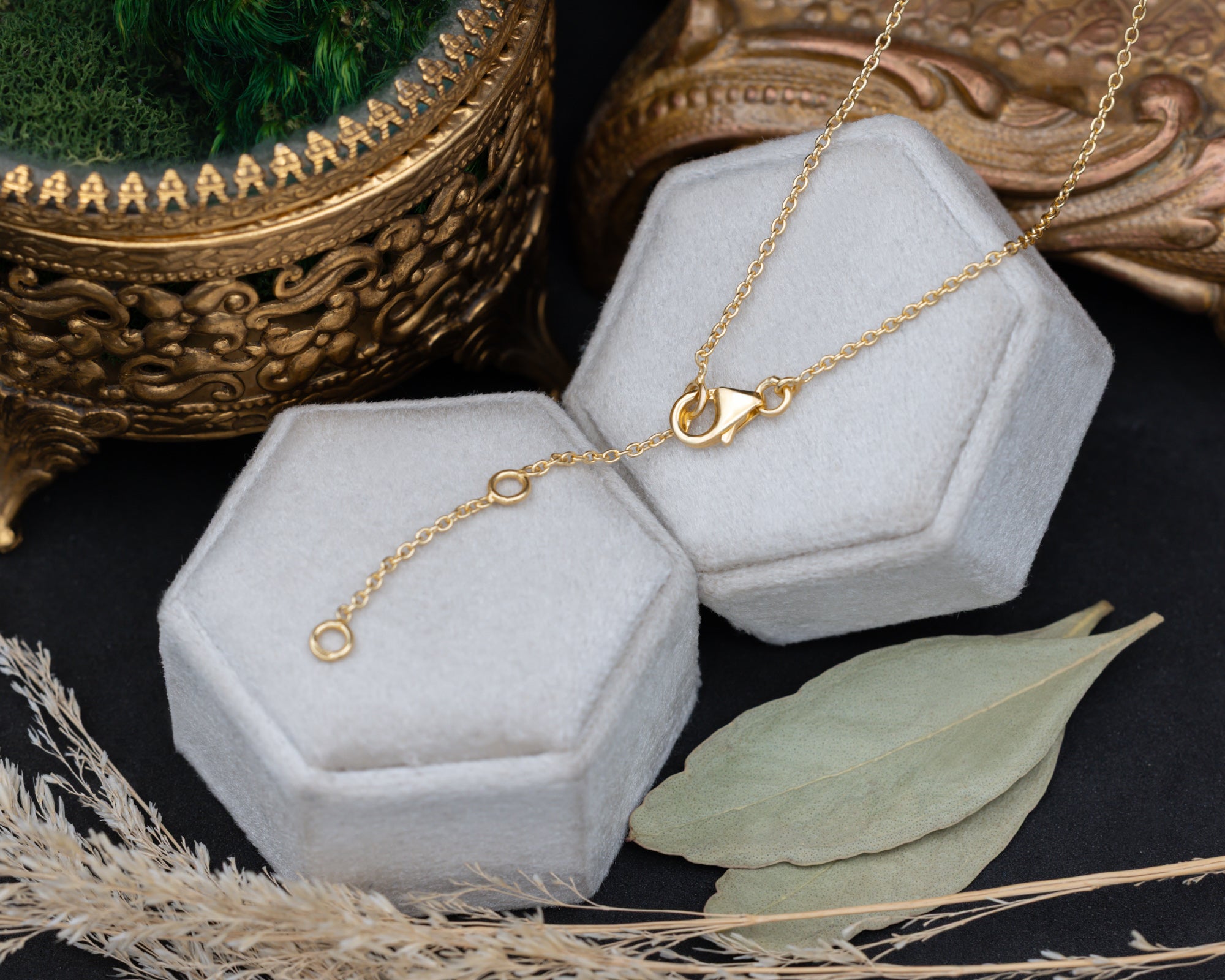 A delicate gold chain necklace with a lobster clasp, draped over two hexagonal white jewelry display boxes on a dark surface, styled with dried leaves and a decorative gold bowl.