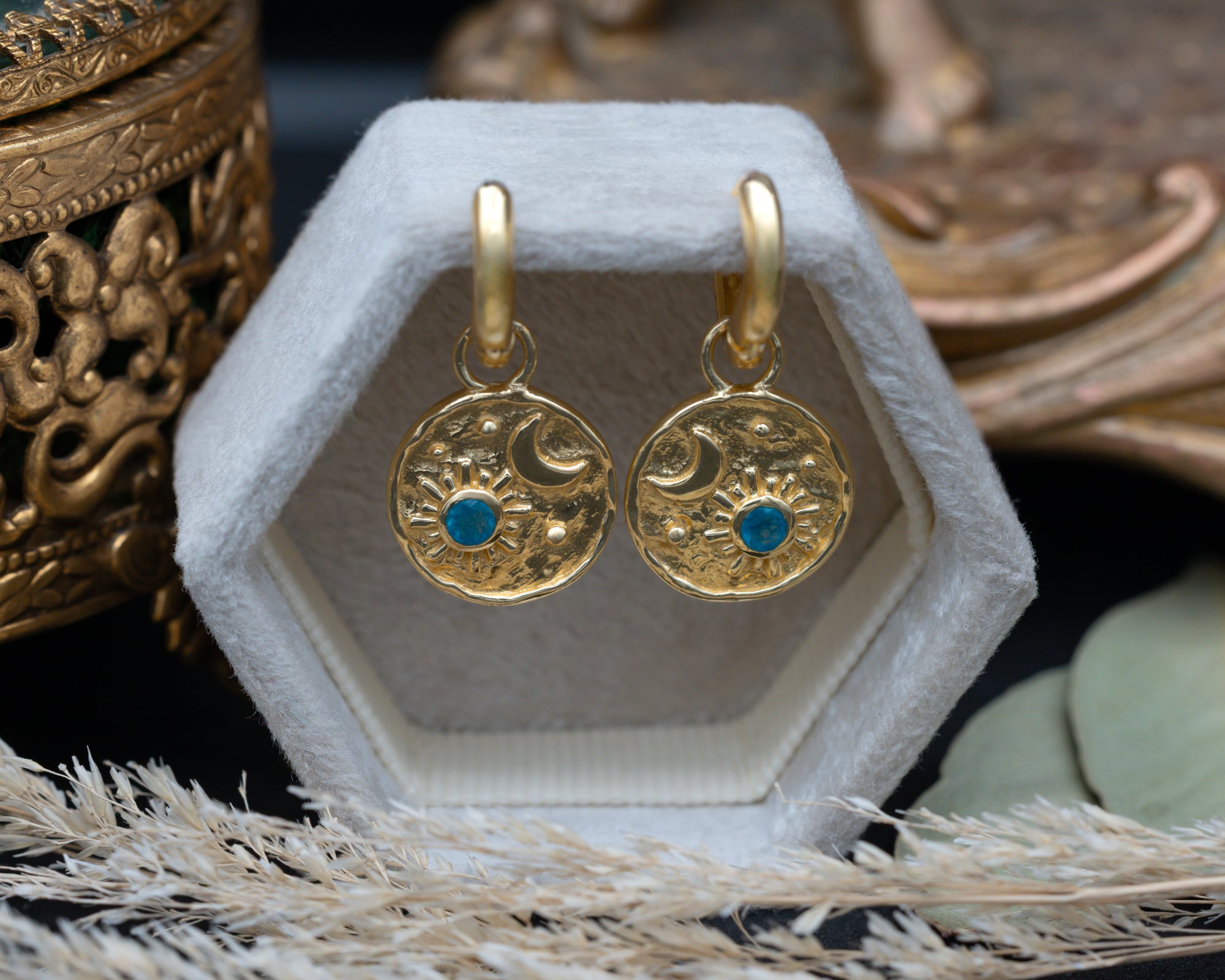 A pair of gold crescent moon and sun earrings with blue apatite gemstone accents , displayed on a velvet jewelry box surrounded by decorative dried grass