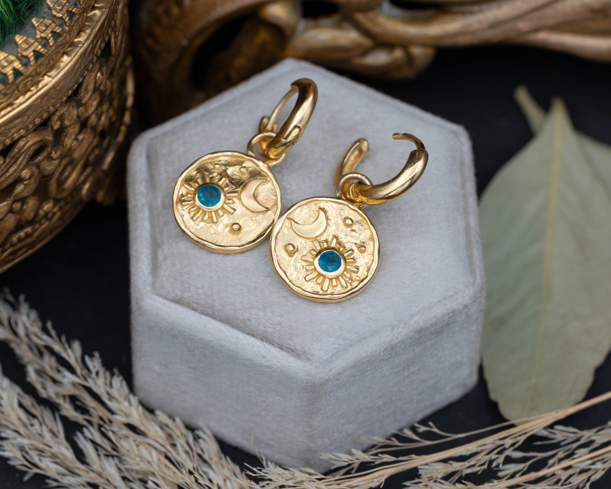 A pair of gold crescent moon and sun earrings with blue apatite gemstone accents , displayed on a velvet jewelry box surrounded by decorative dried grass