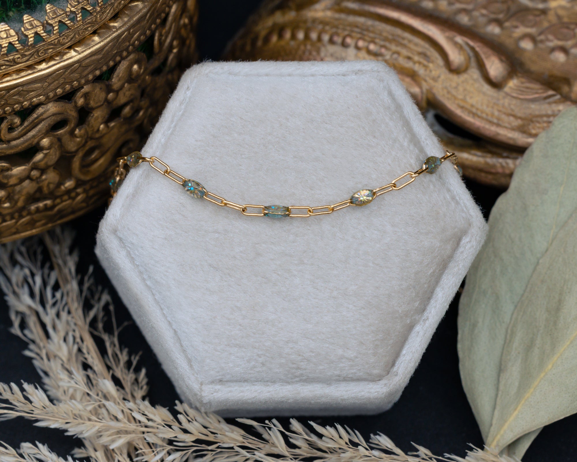 A gold chain necklace with multi-color beads draped over a hexagonal jewelry box, styled with dried grass and leaves on a dark surface.