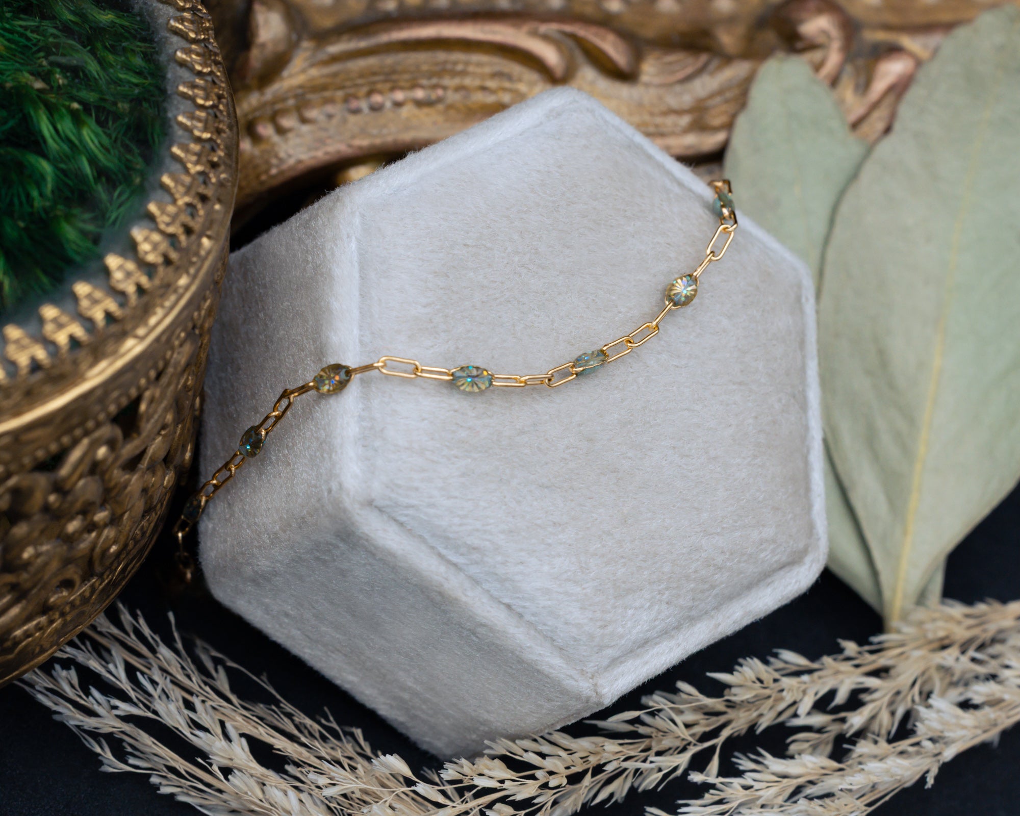 A gold chain necklace with multi-color beads draped over a hexagonal jewelry box, styled with dried grass and leaves on a dark surface.