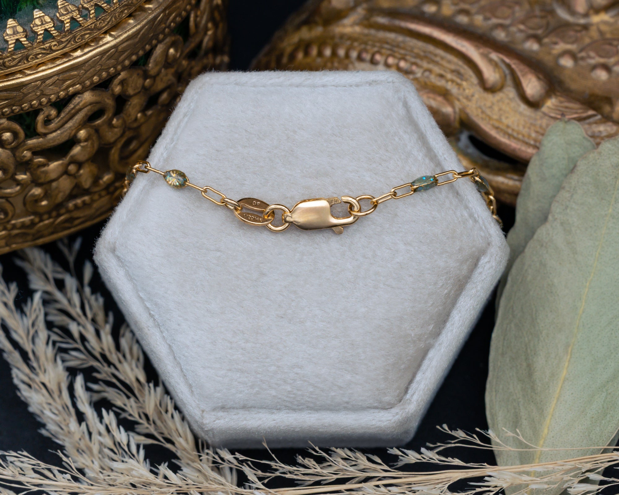 A gold chain necklace with lobster clasp draped over a hexagonal jewelry box, styled with dried grass and leaves on a dark surface.