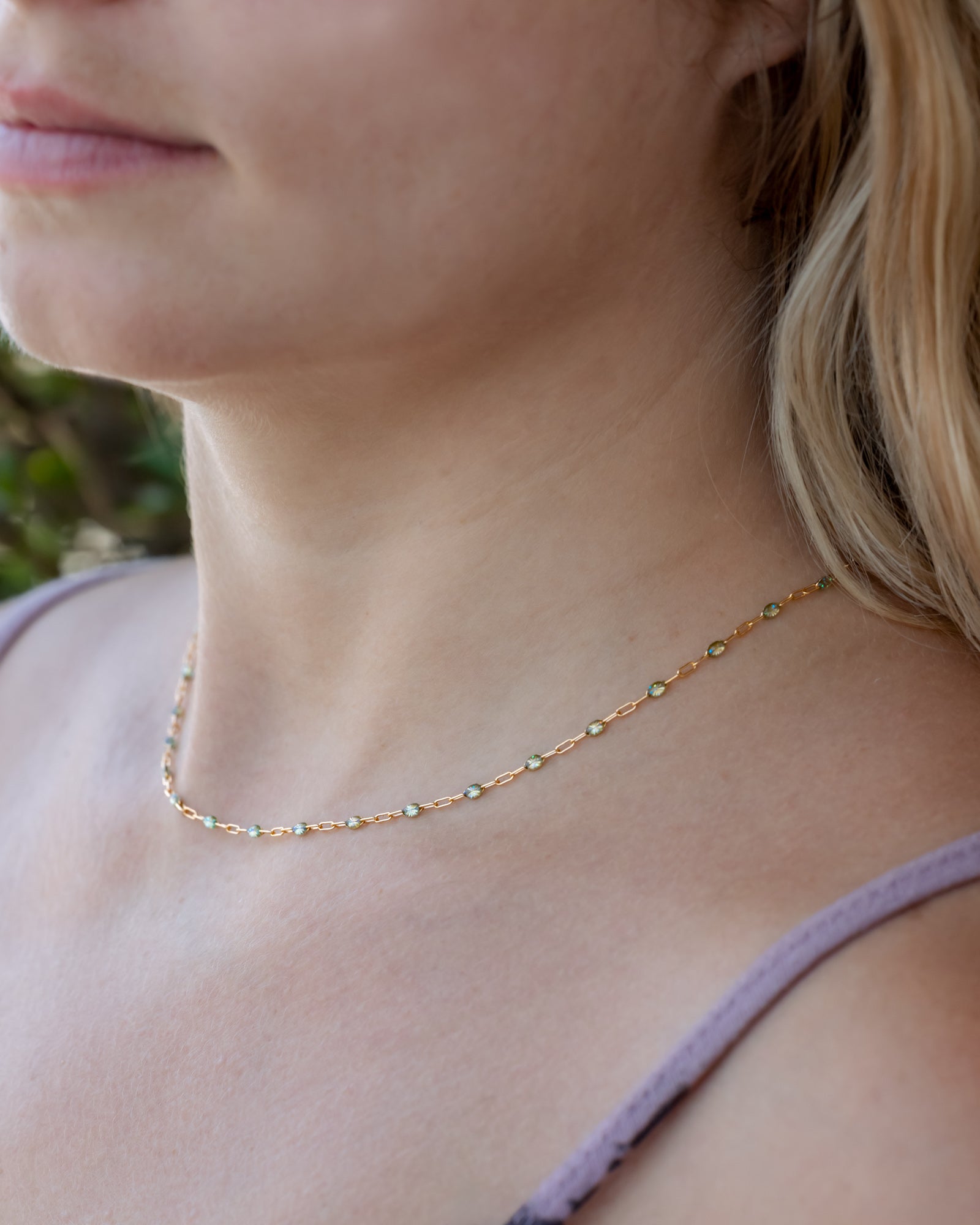 A close-up of a person with blond hair wearing a delicate gold chain necklace with multi-colored beads.