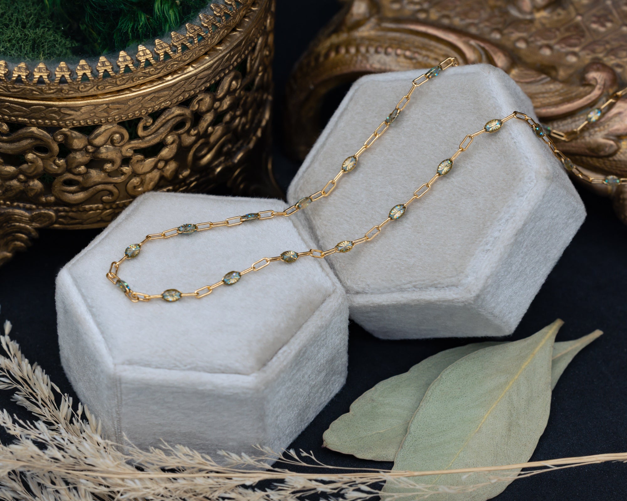 A gold chain necklace with multi-color beads draped over two hexagonal jewelry boxes, styled with dried grass and leaves on a dark surface.