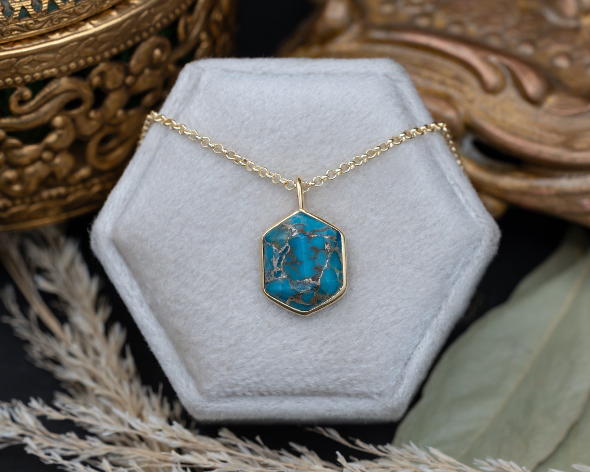 A turquoise pendant on a gold chain against a white cushion with dried flowers in the background