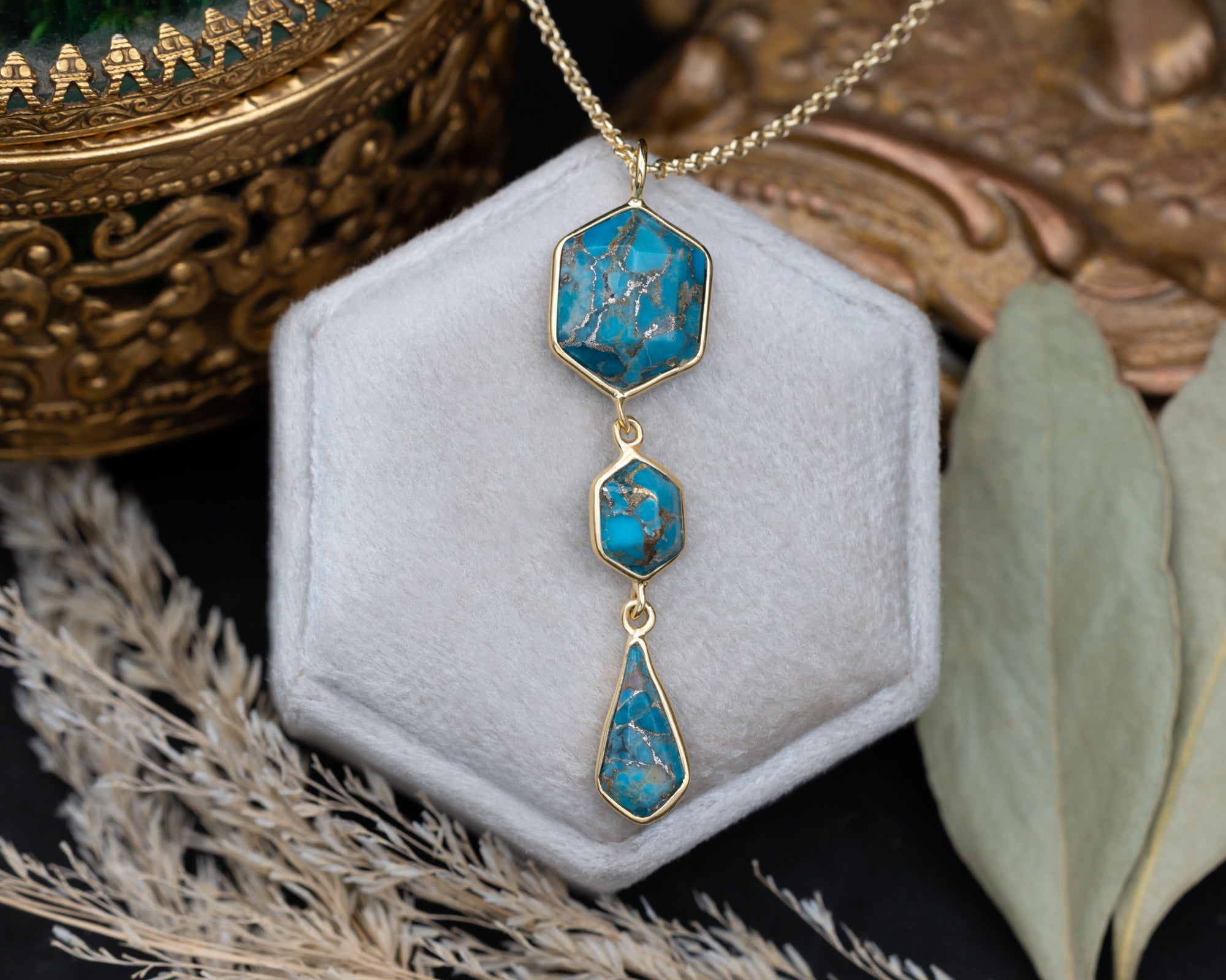 A gold pendant necklace with three turquoise stones on a gold chain, sitting on a white cushion with dried flowers in the background