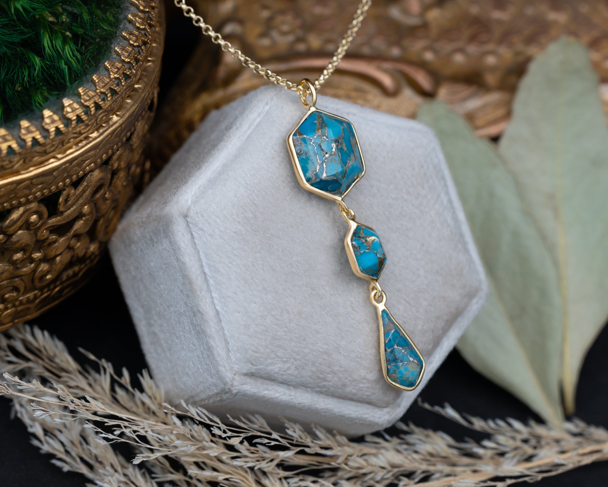 A gold pendant necklace with three turquoise stones on a gold chain, sitting on a white cushion with dried flowers in the background
