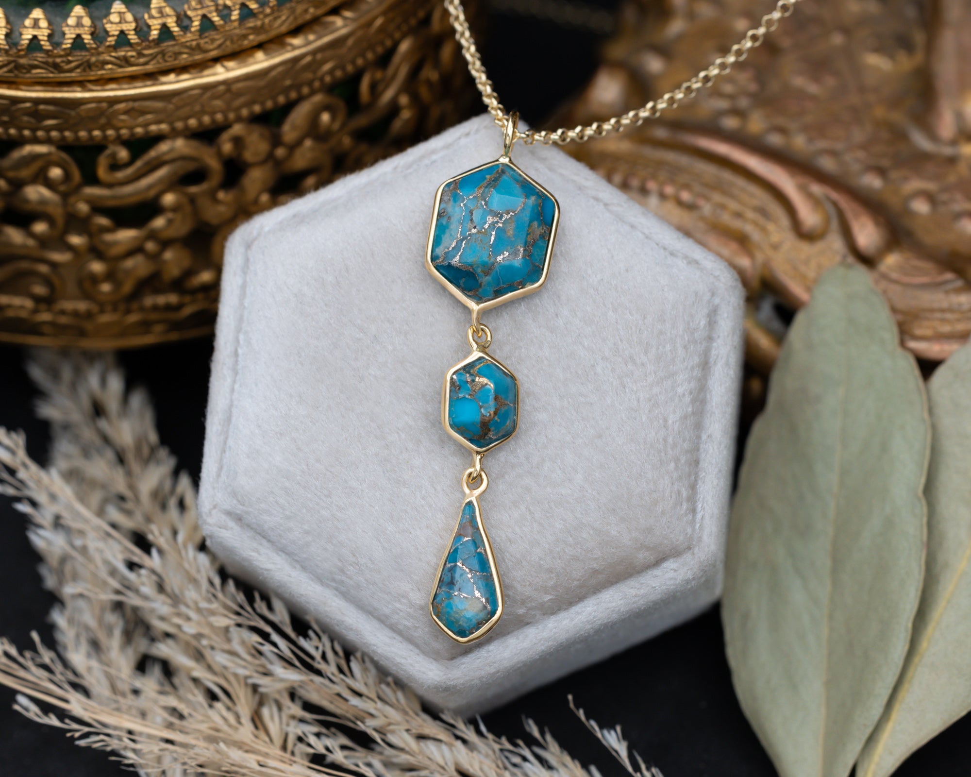 A gold pendant necklace with three turquoise stones on a gold chain, sitting on a white cushion with dried flowers in the background