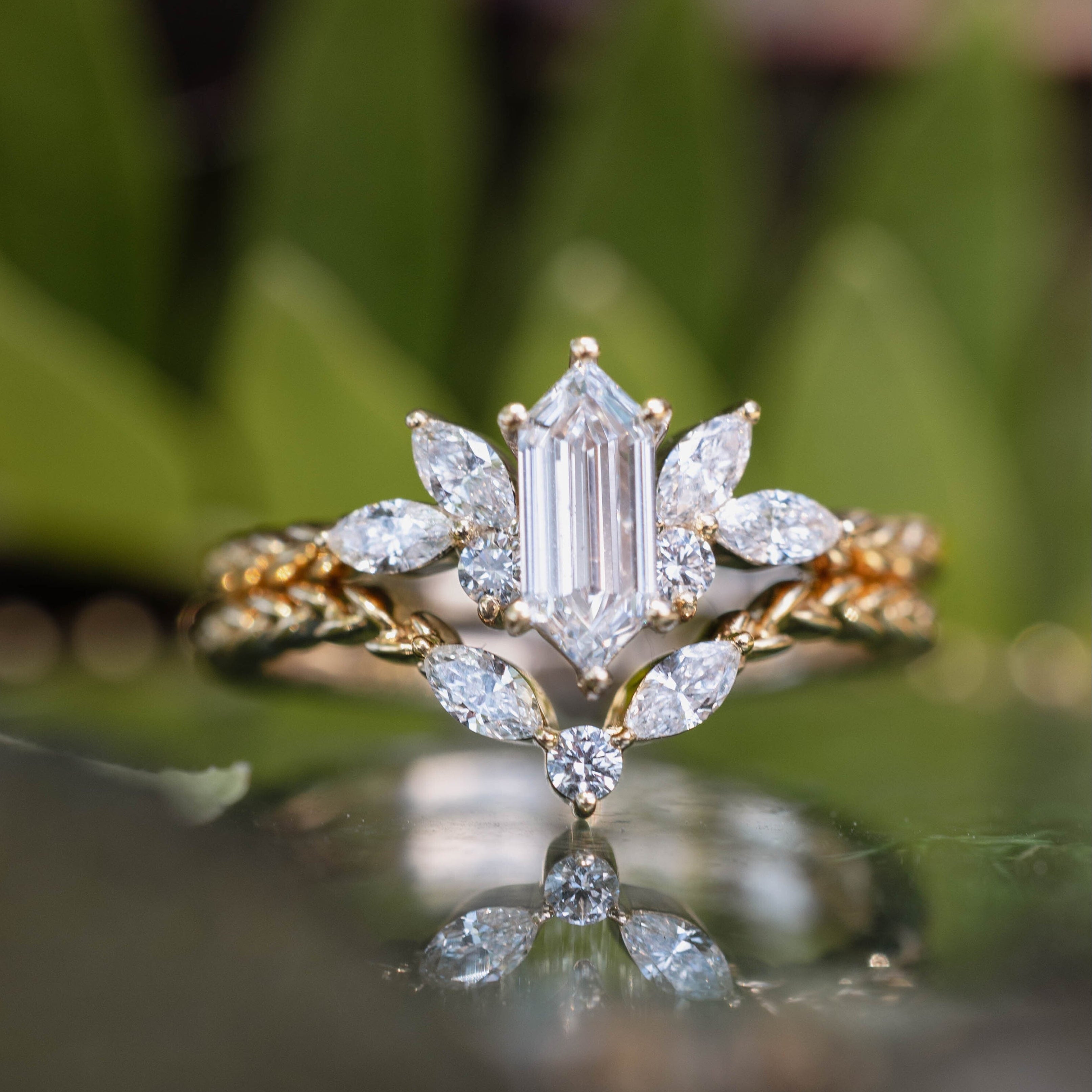 Gold ring with diamond center stone and diamond tracer on a reflective surface with green foliage in the background