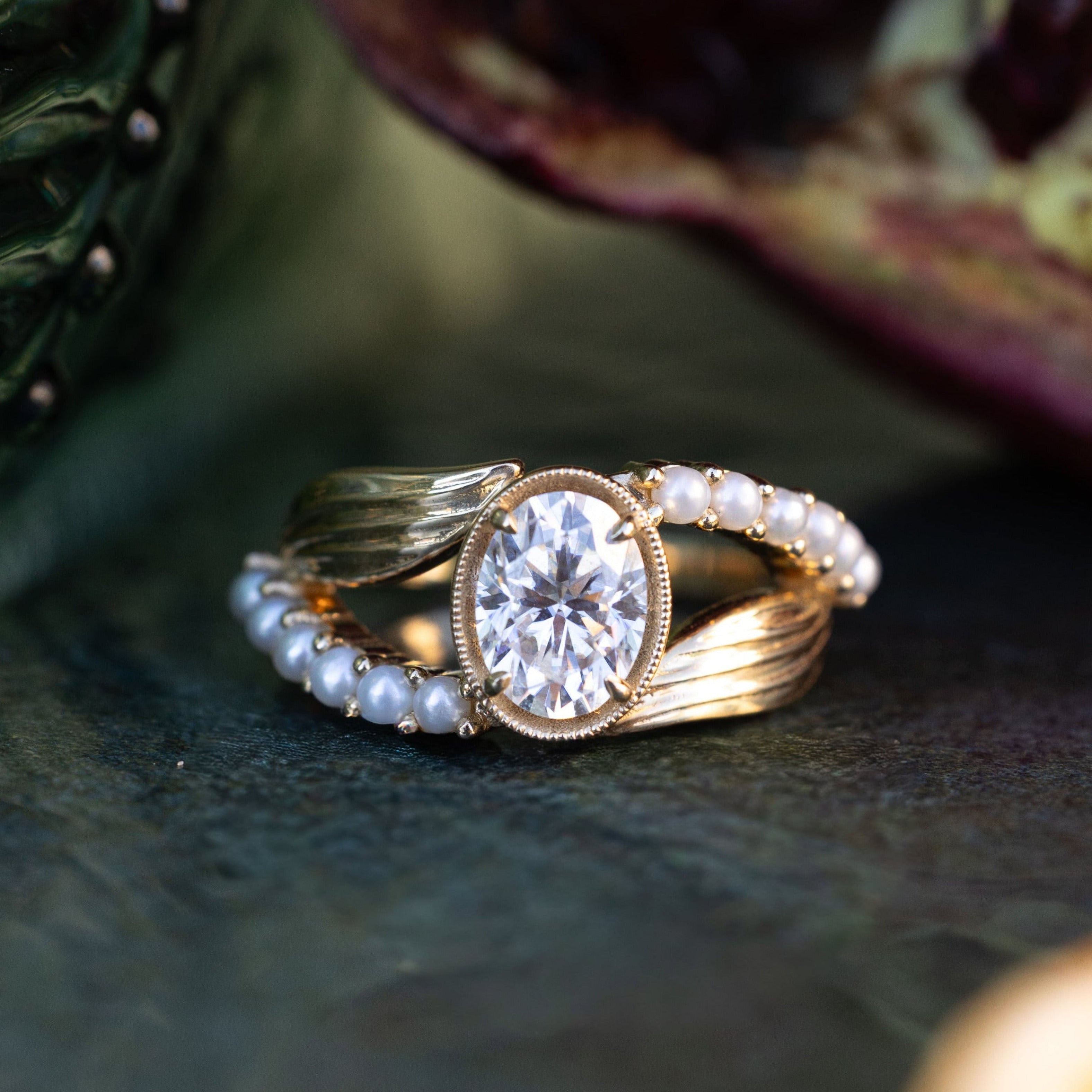 Gold ring with a moissanite center stone on a textured surface with pomegranate in the background