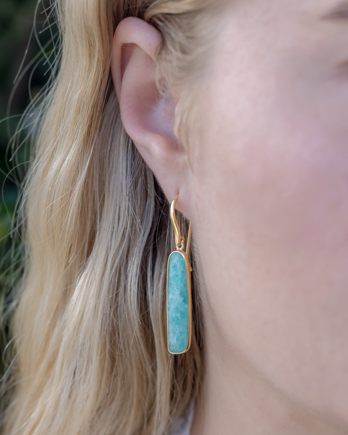 Person with blond hair wearing gold dangle earring with oblong amazonite gemstone
