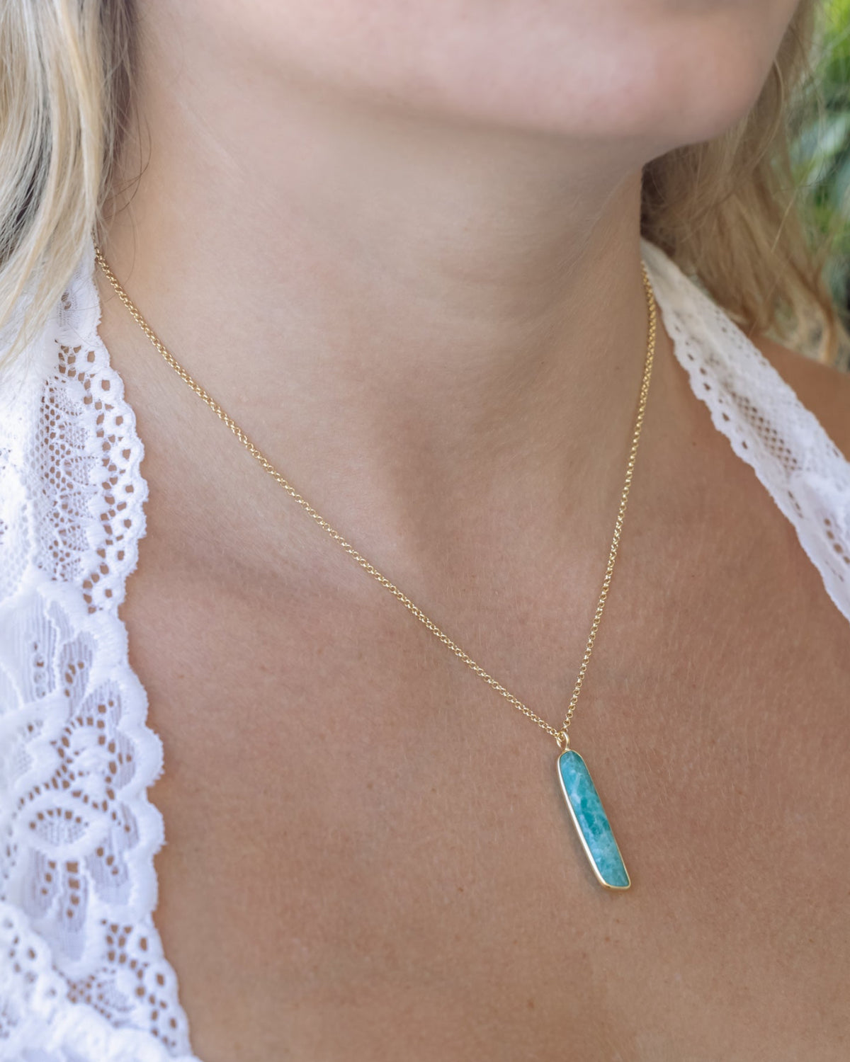 Person wearing gold necklace with oblong amazonite gemstone pendant against a leafy background
