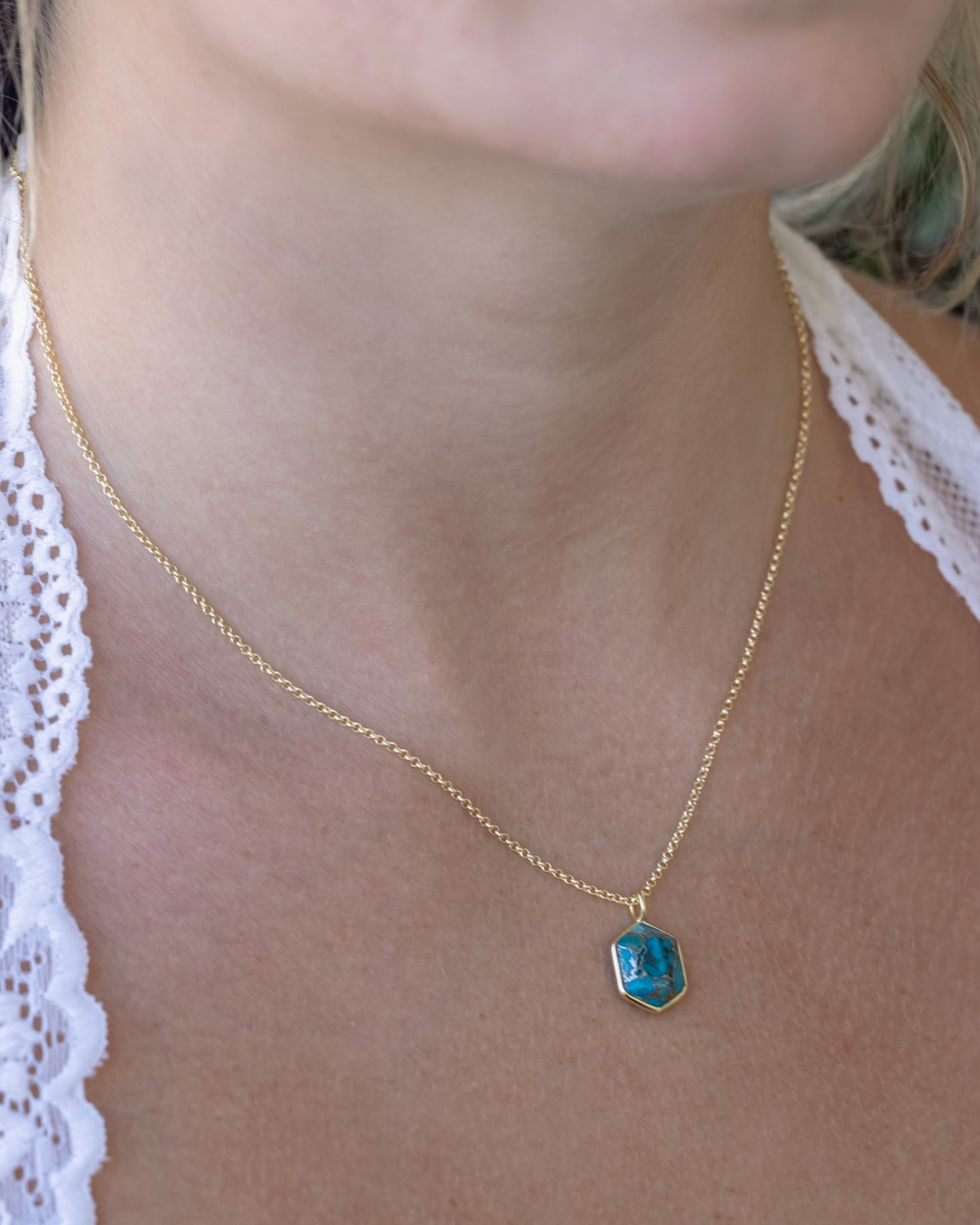 Person wearing gold necklace with hexagon cut turquoise pendant against a leafy background