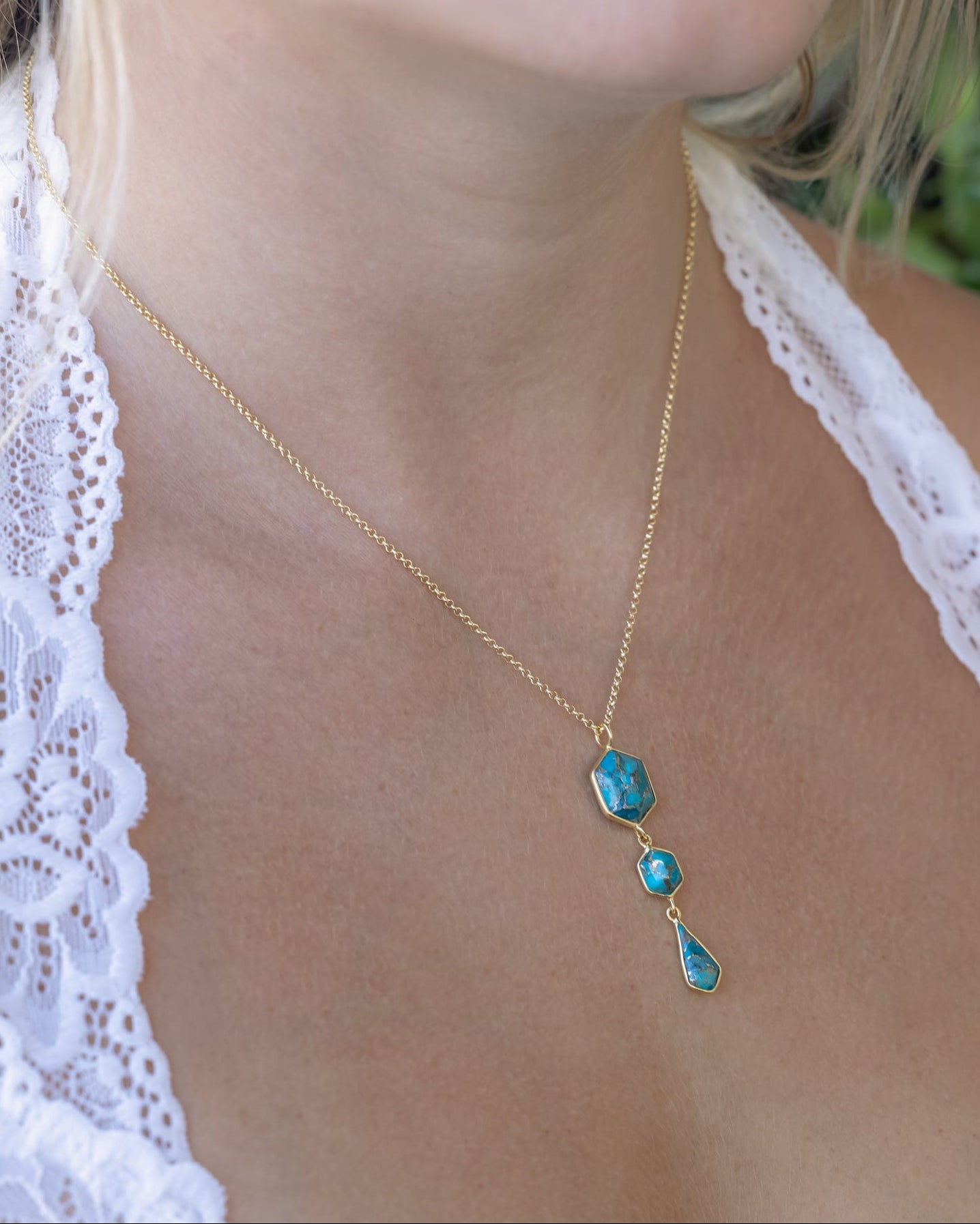 Person wearing gold necklace with triple turquoise dangle pendant against a leafy background