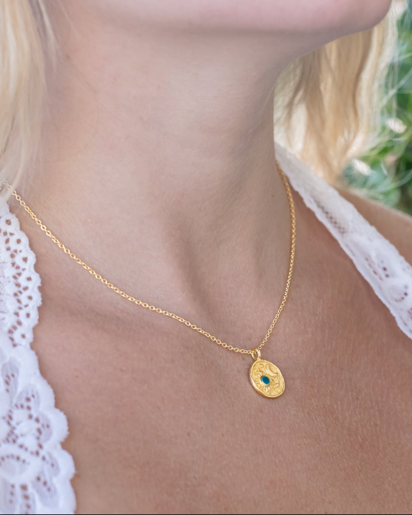 Person wearing gold necklace on their neck with sun and moon design with apatite accent against a leafy background
