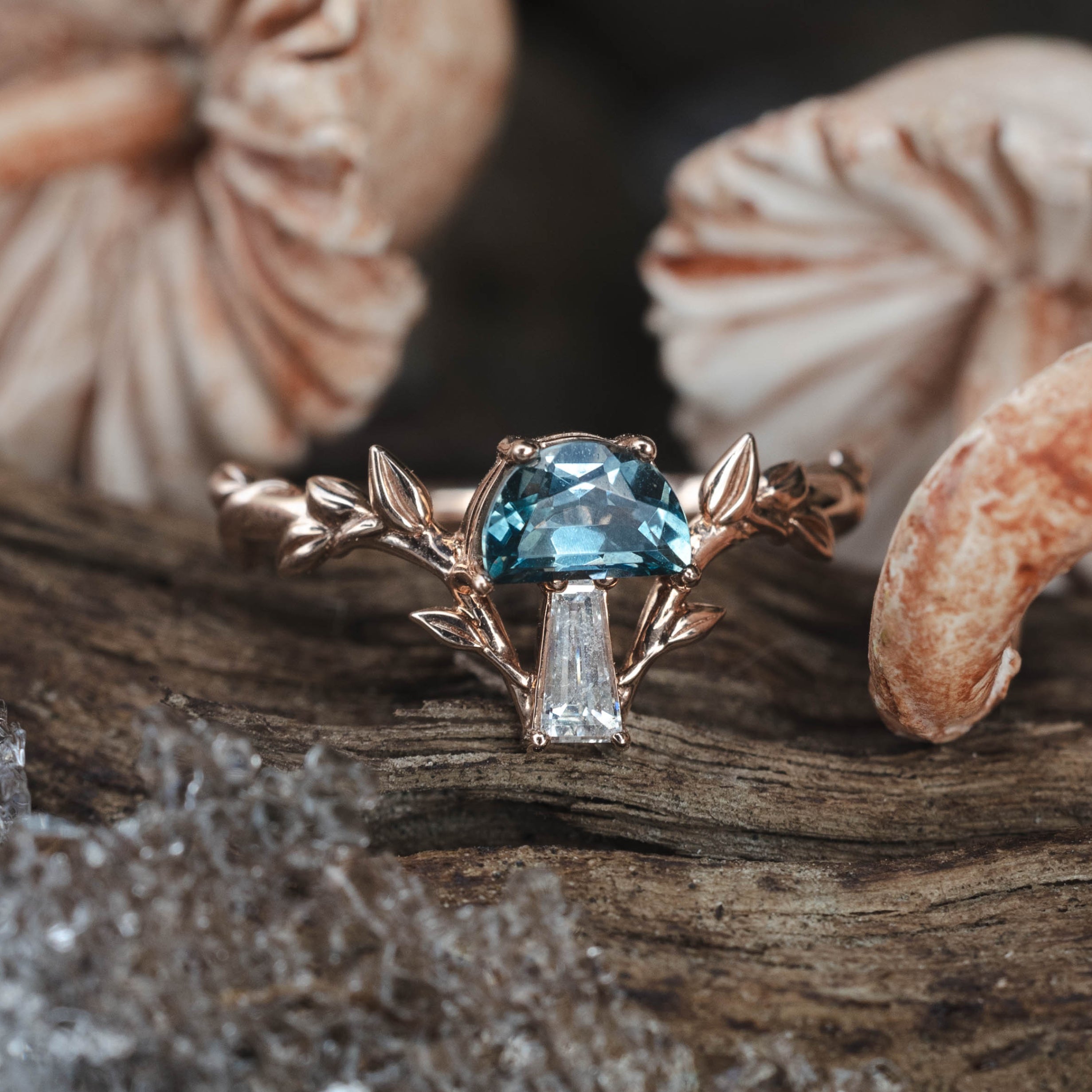 Rose gold ring with blue gemstone and diamond on a wooden surface with mushrooms in the background