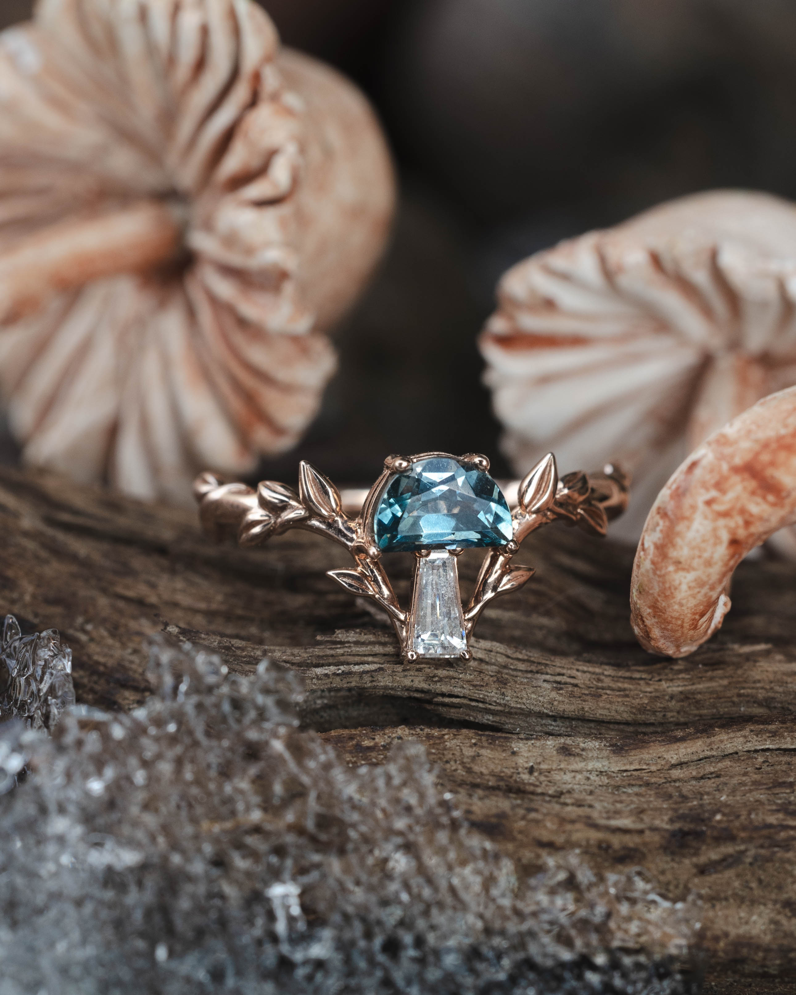 Rose gold ring with blue gemstone and diamond on a wooden surface with mushrooms in the background