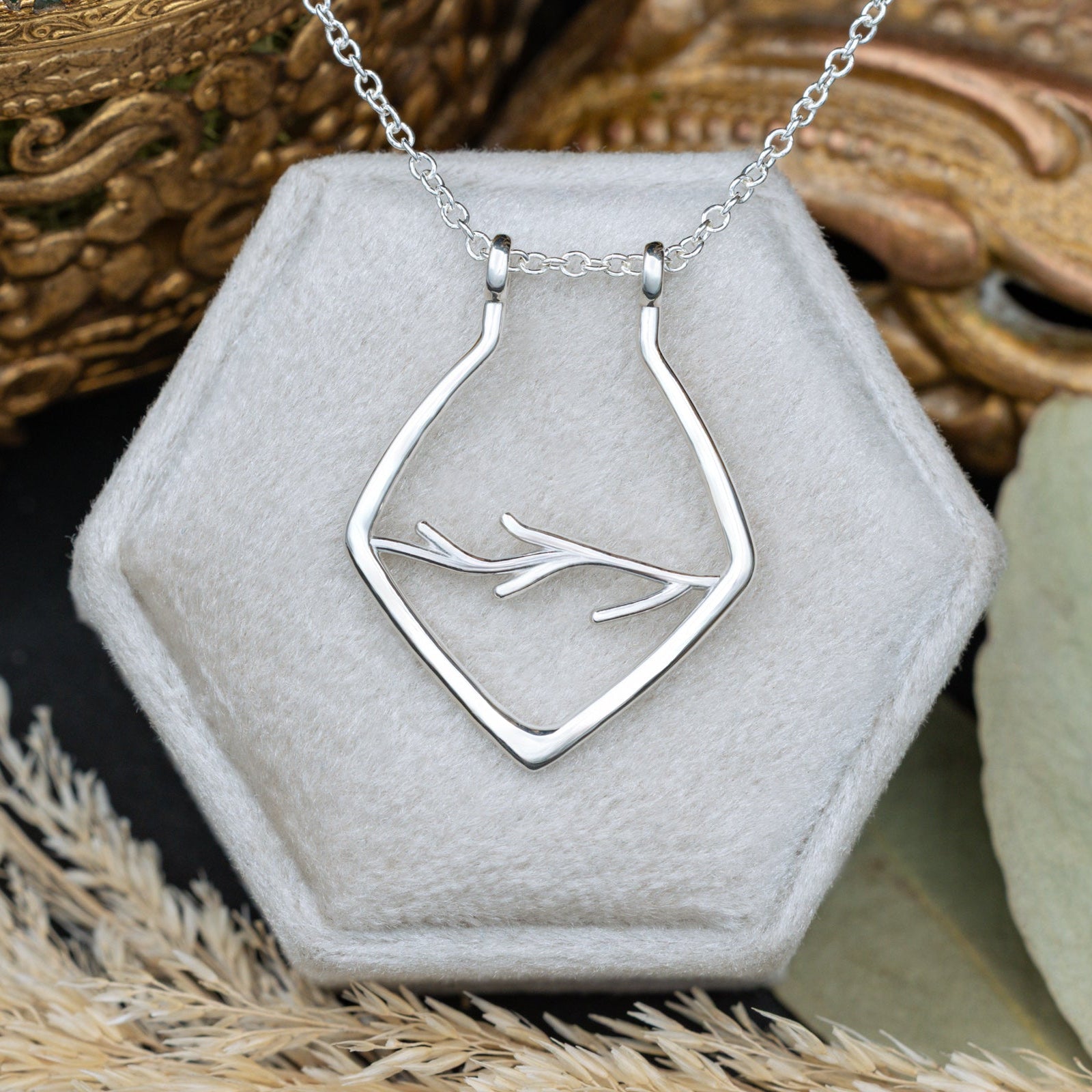 A silver necklace with antler design on a white cushion with blurred dried flowers