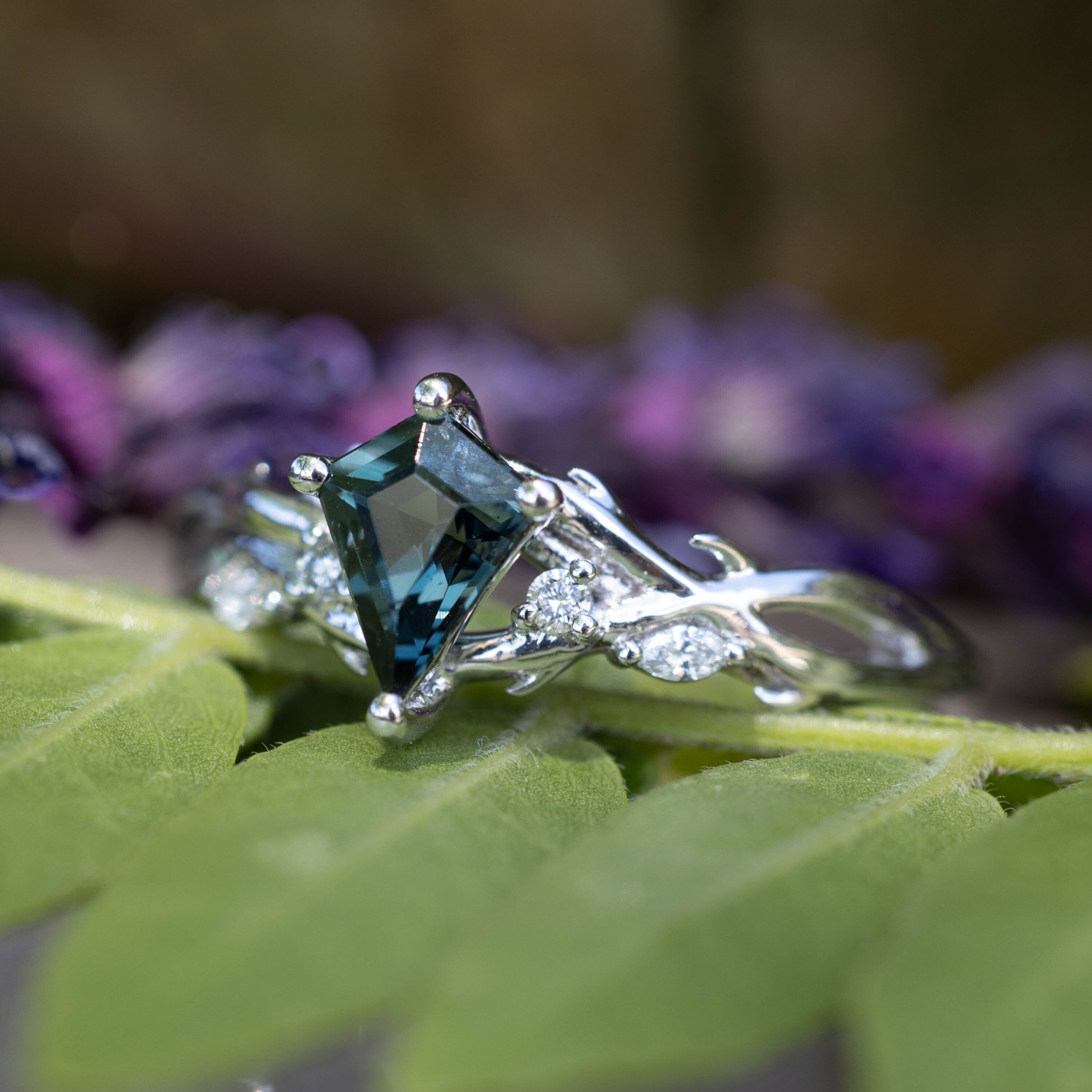 White gold ring with a blue spinel on a leaf with blurred purple flowers in the background