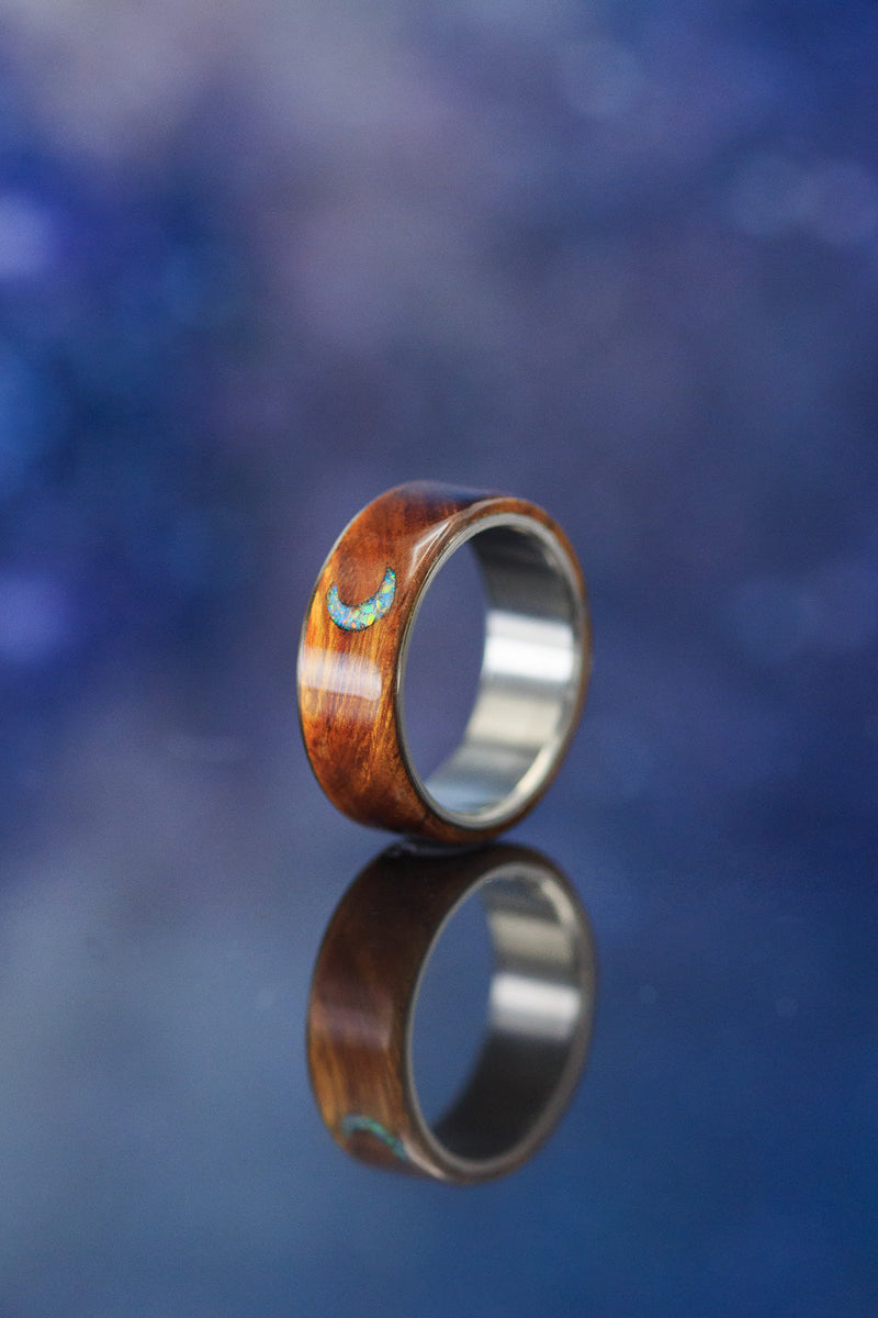 Shown here is  The "Theron", a custom, handcrafted men's wedding ring featuring an ironwood overlay that has a crescent moon engraved into it. The moon itself is filled with white crushed fire and ice opal. Additional inlay options are available upon request.